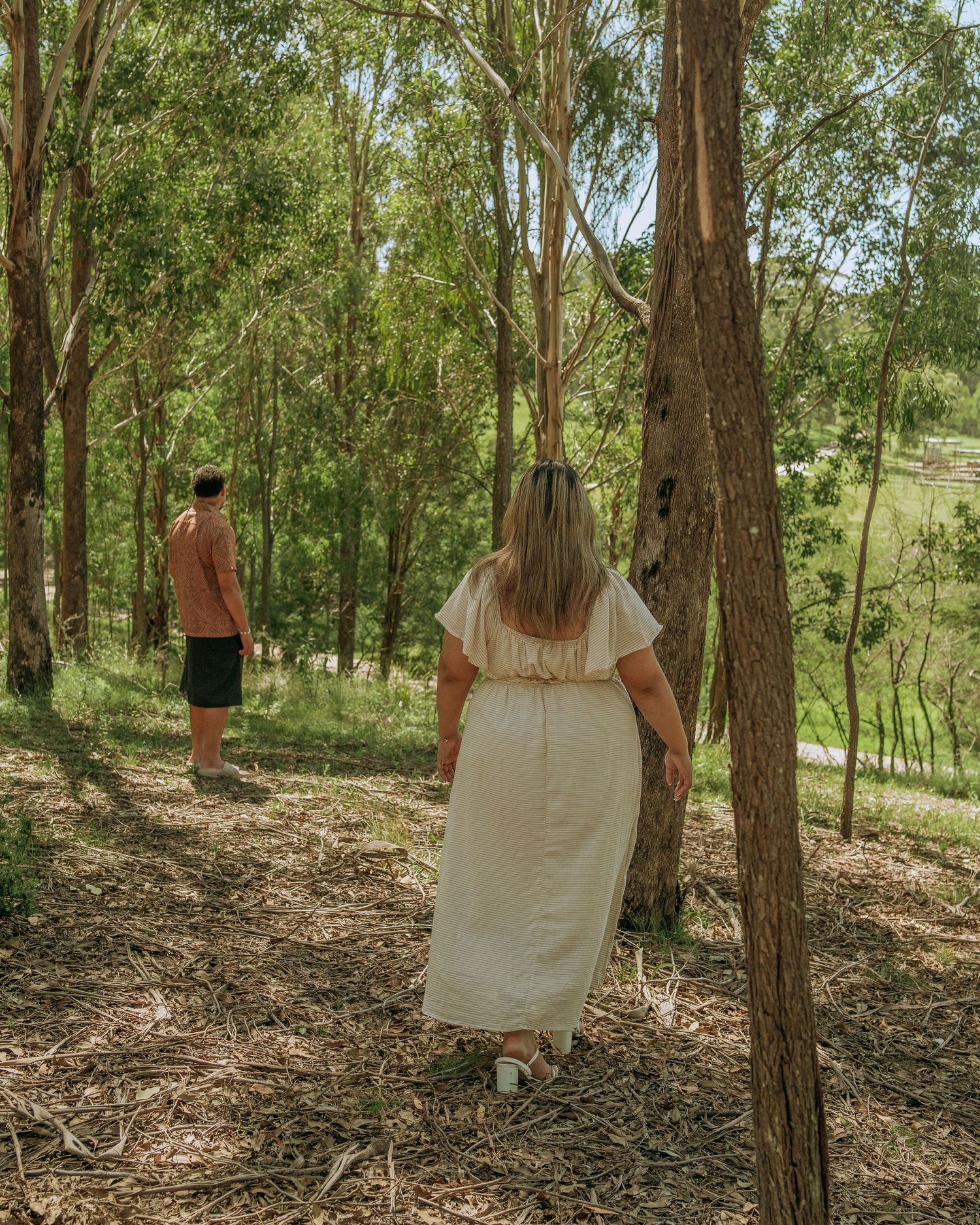 Couple walking separately through a sunlit eucalyptus forest toward a scenic green valley below.