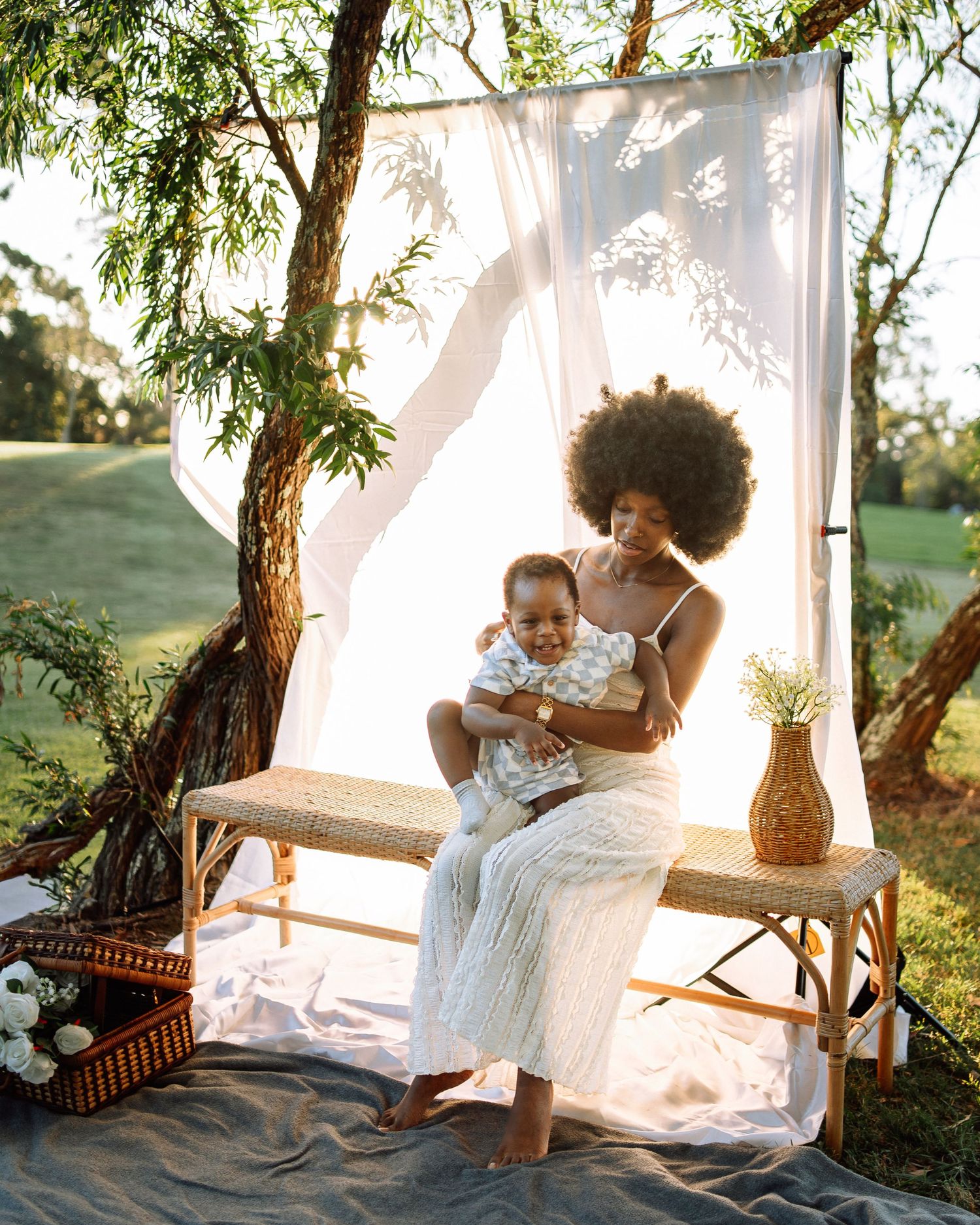 Black mother with afro sitting on bench holding baby, posed before sheer white backdrop outdoors.