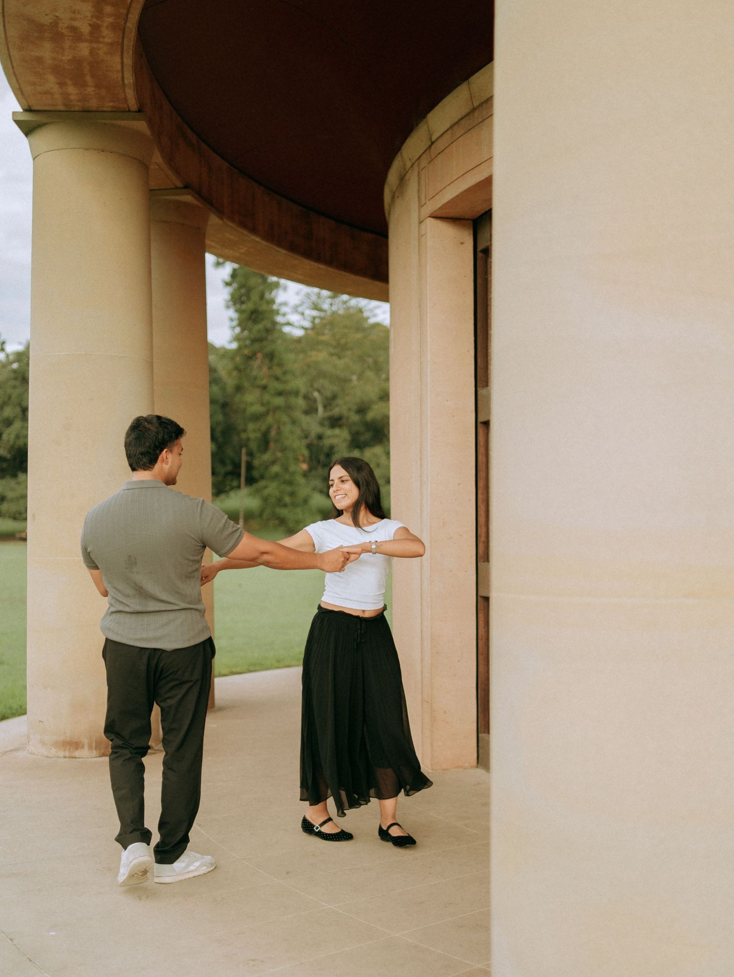 A couple dances playfully under a curved architectural portico with lush green gardens in the background.