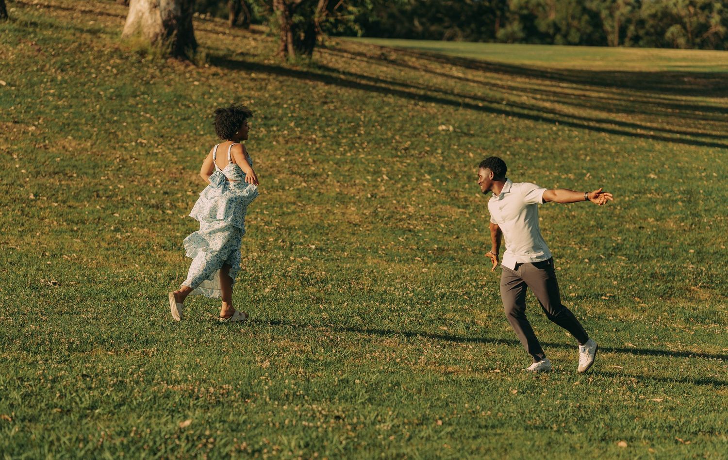 A playful couple chases each other across a sunlit grassy park surrounded by autumn trees.