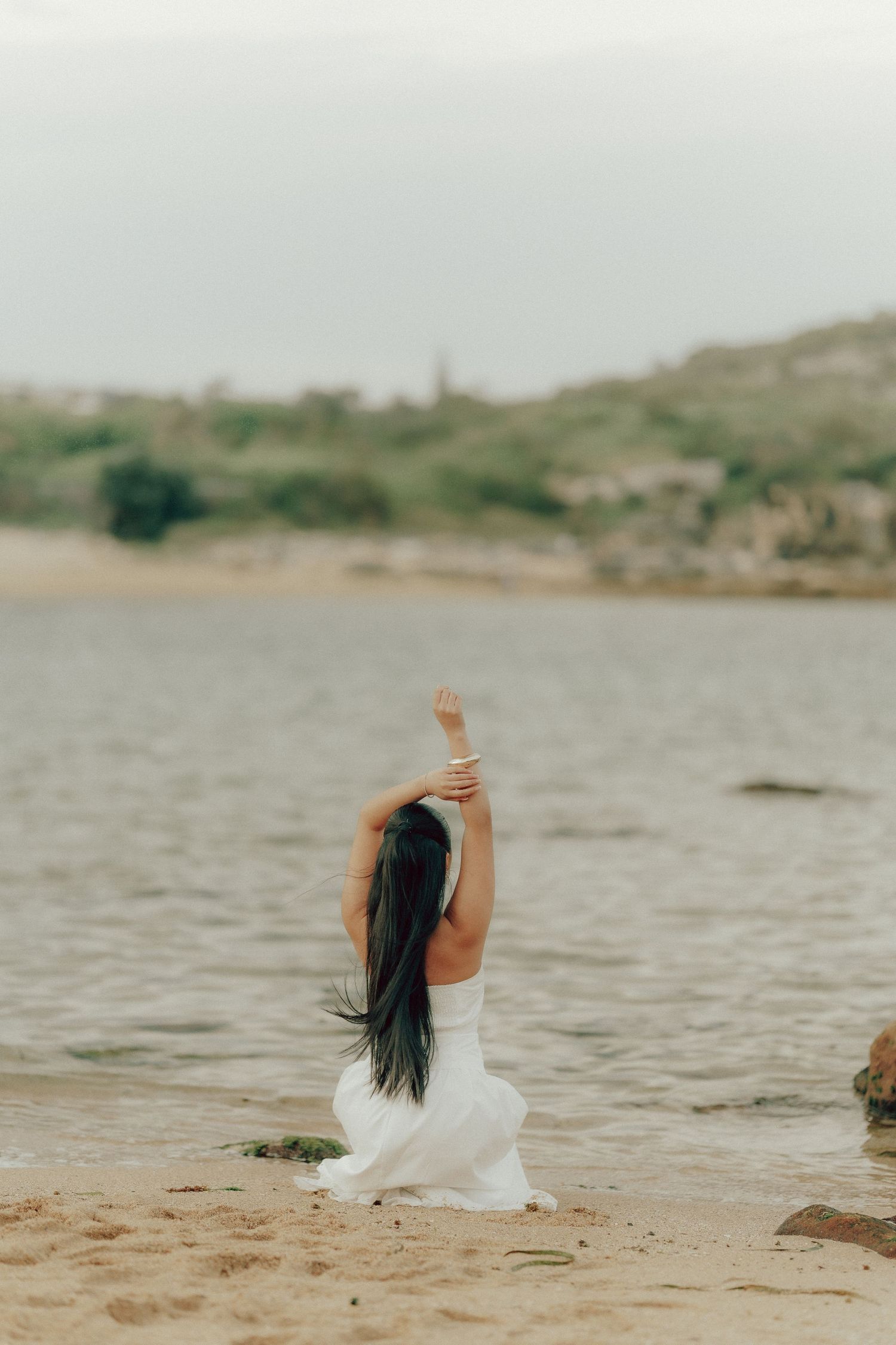 Woman in white dress kneeling on beach sand with arm raised toward sky near coastal waters.