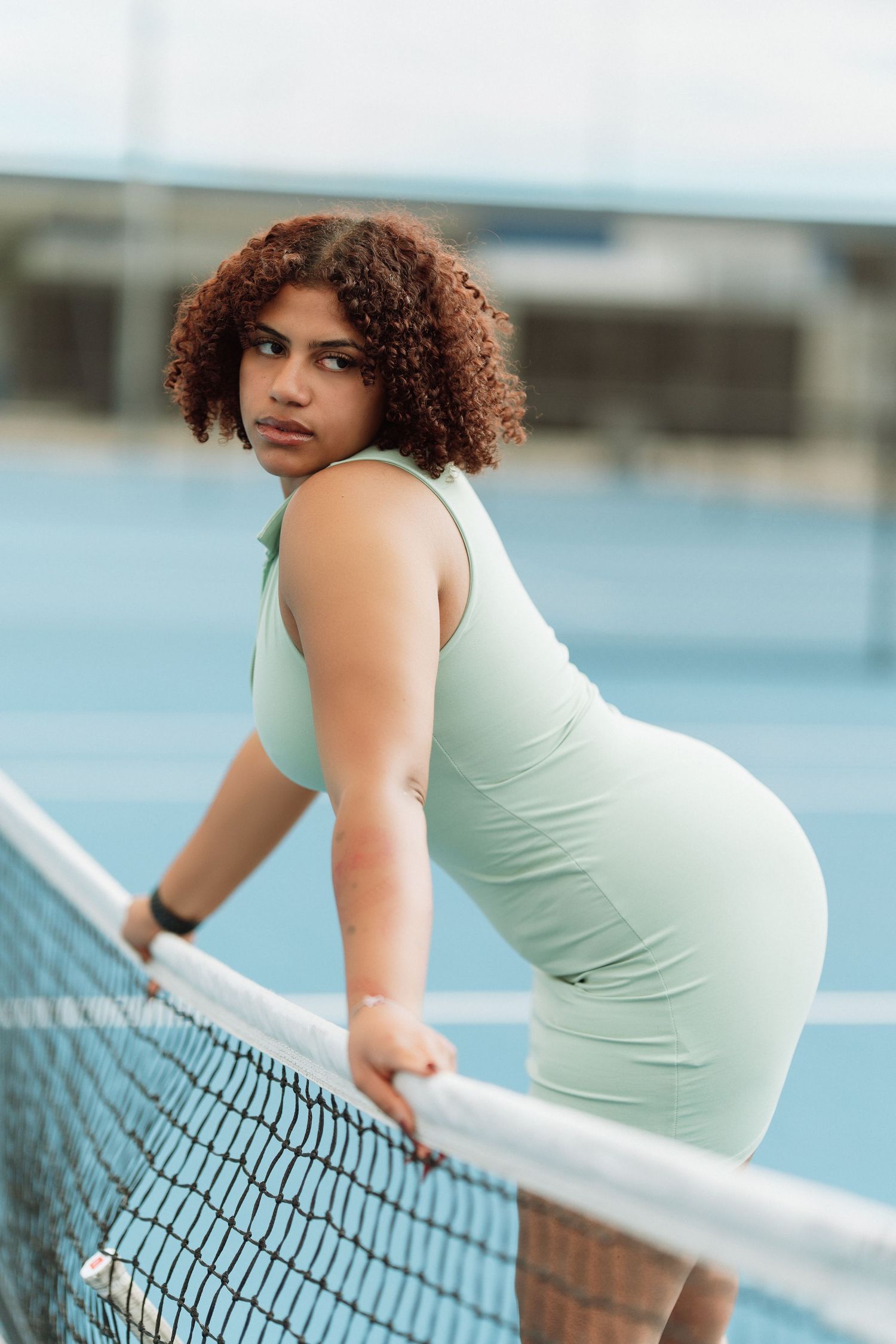 A woman in a fitted mint green tennis dress leans on the court net, looking back at the camera confidently.