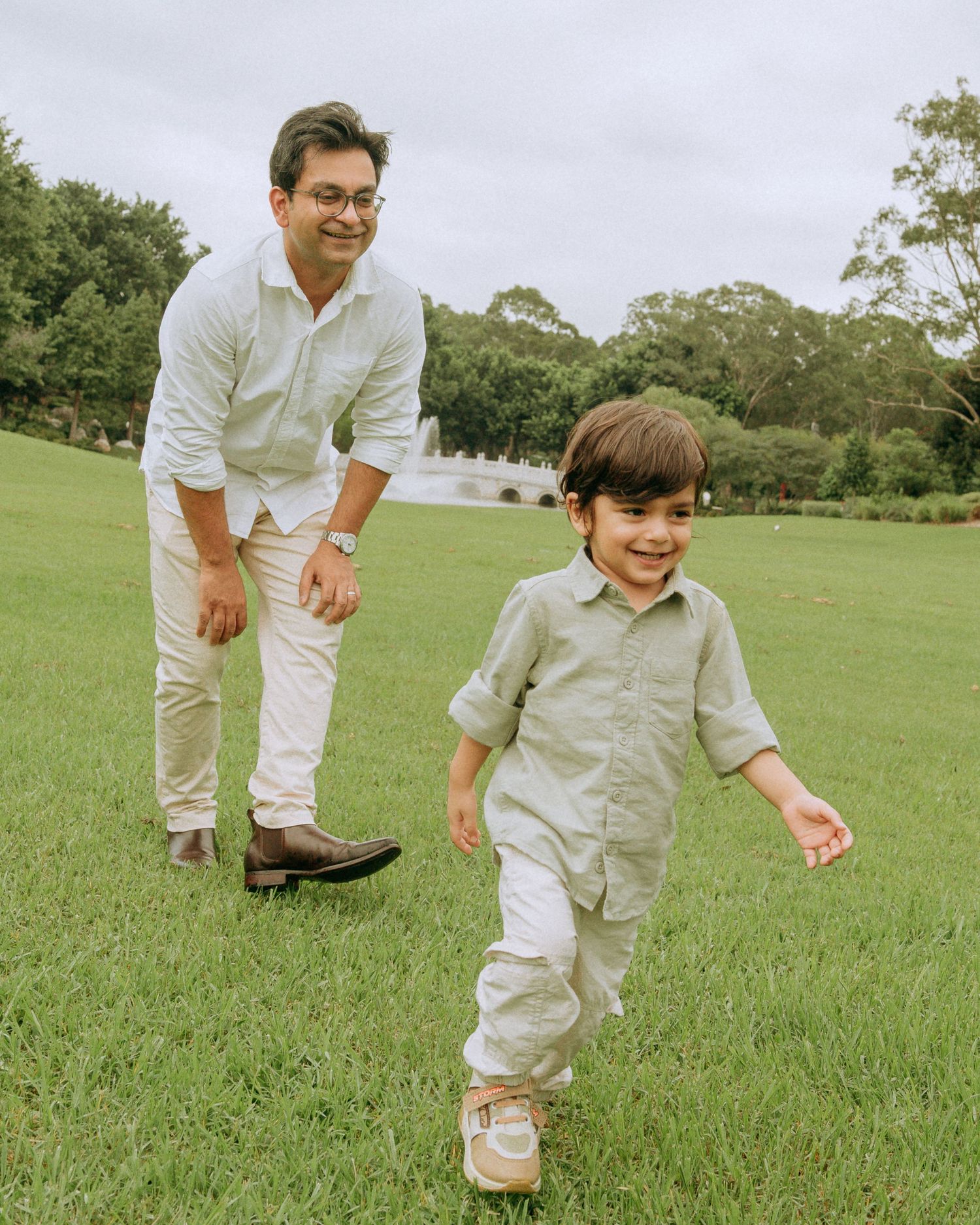 A father smiles proudly as his young son runs ahead of him on a lush green lawn outdoors.