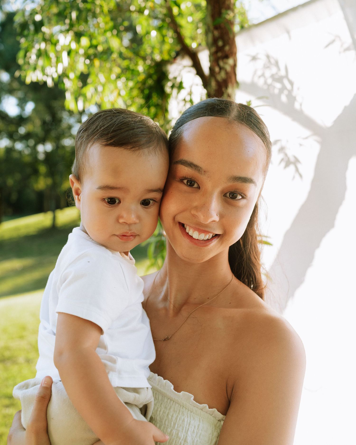 Beautiful young mother smiling while holding cute baby boy cheek-to-cheek in bright outdoor natural light portrait.