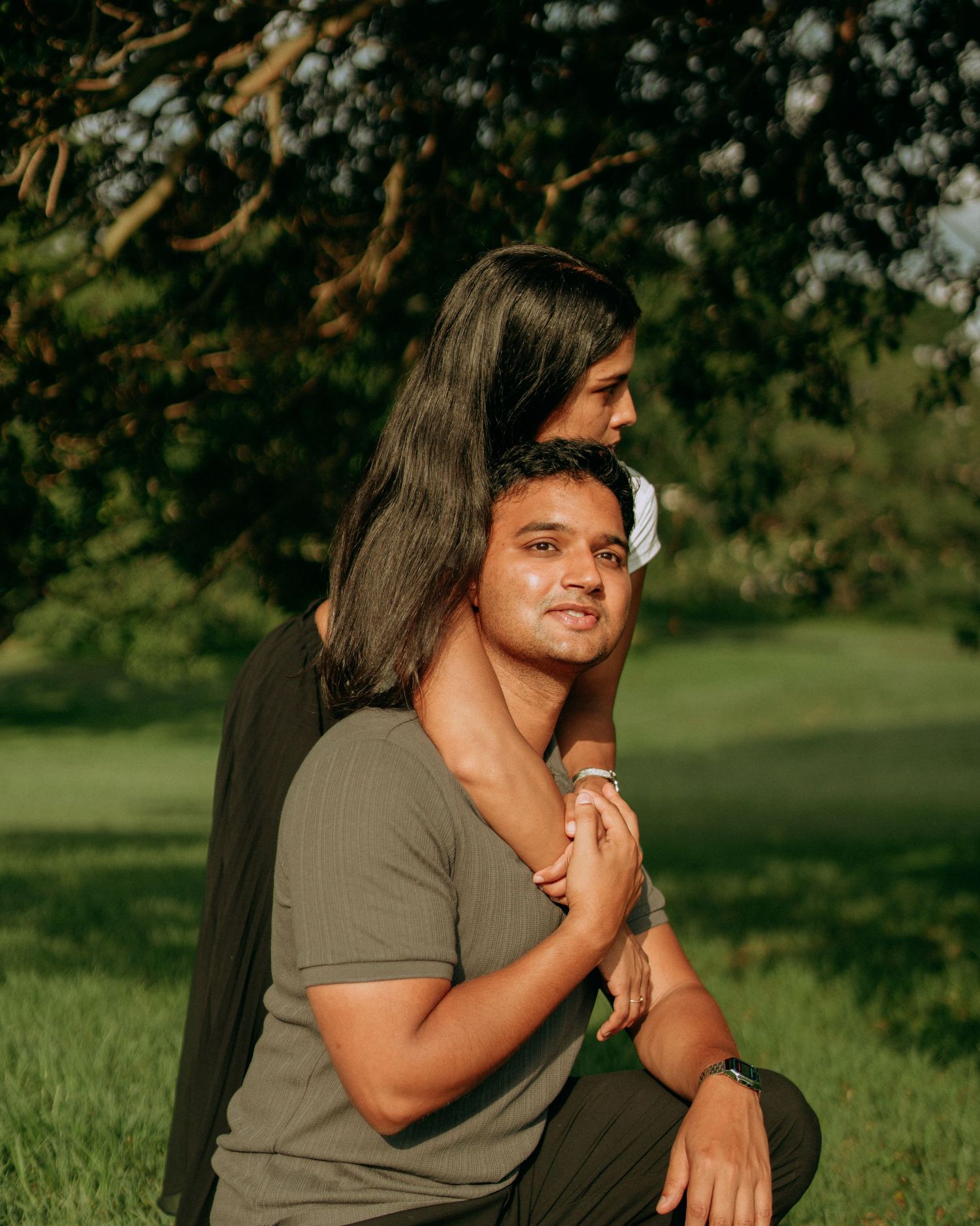 A young man sits outdoors while his partner embraces him from behind under a shady green tree.