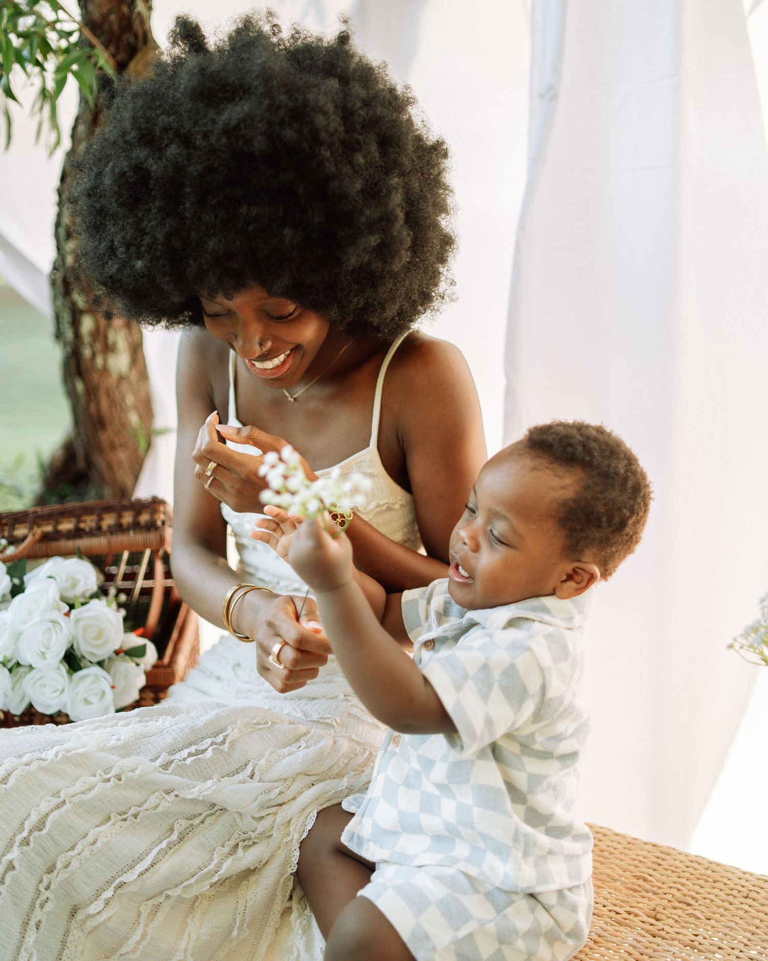 Black mother with afro smiling as her toddler son reaches for flowers in a bright outdoor setting.