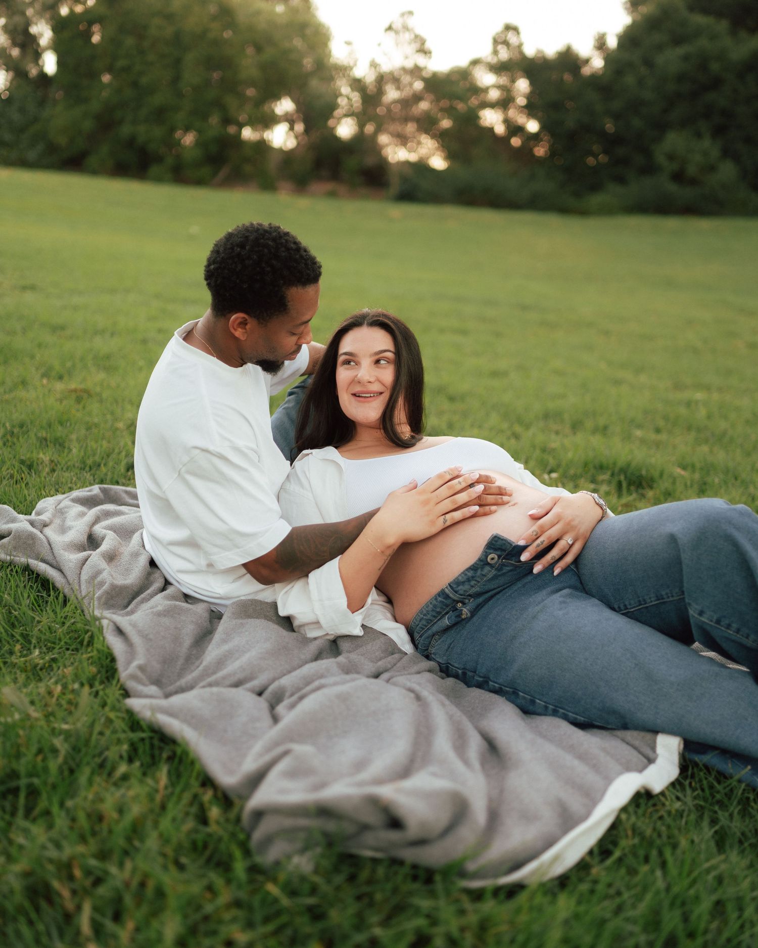Couple relaxing on a blanket in a green park, gently cradling the pregnant woman's bare belly.