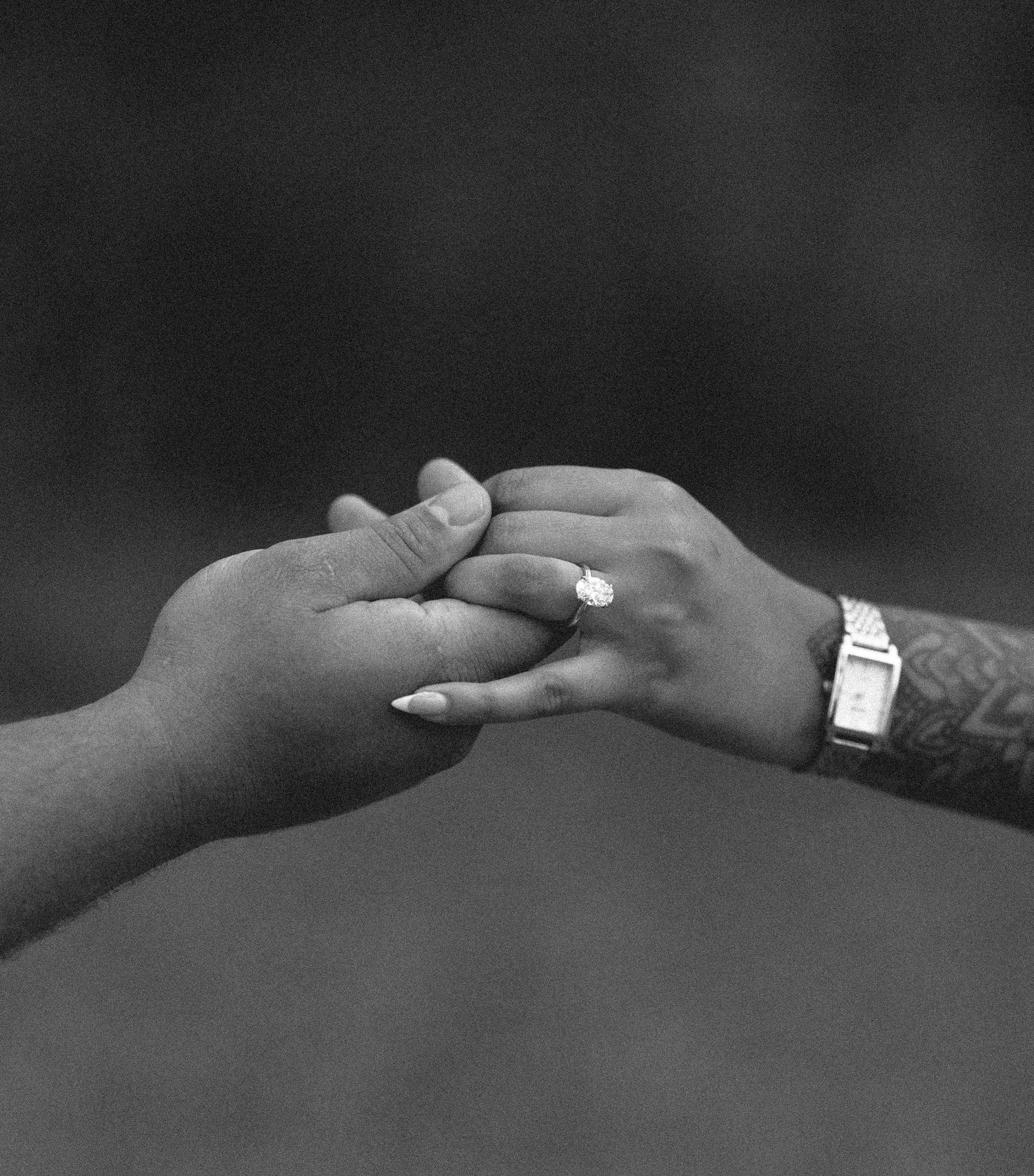 A black and white close-up of two hands holding, showcasing a diamond engagement ring and tattoos.