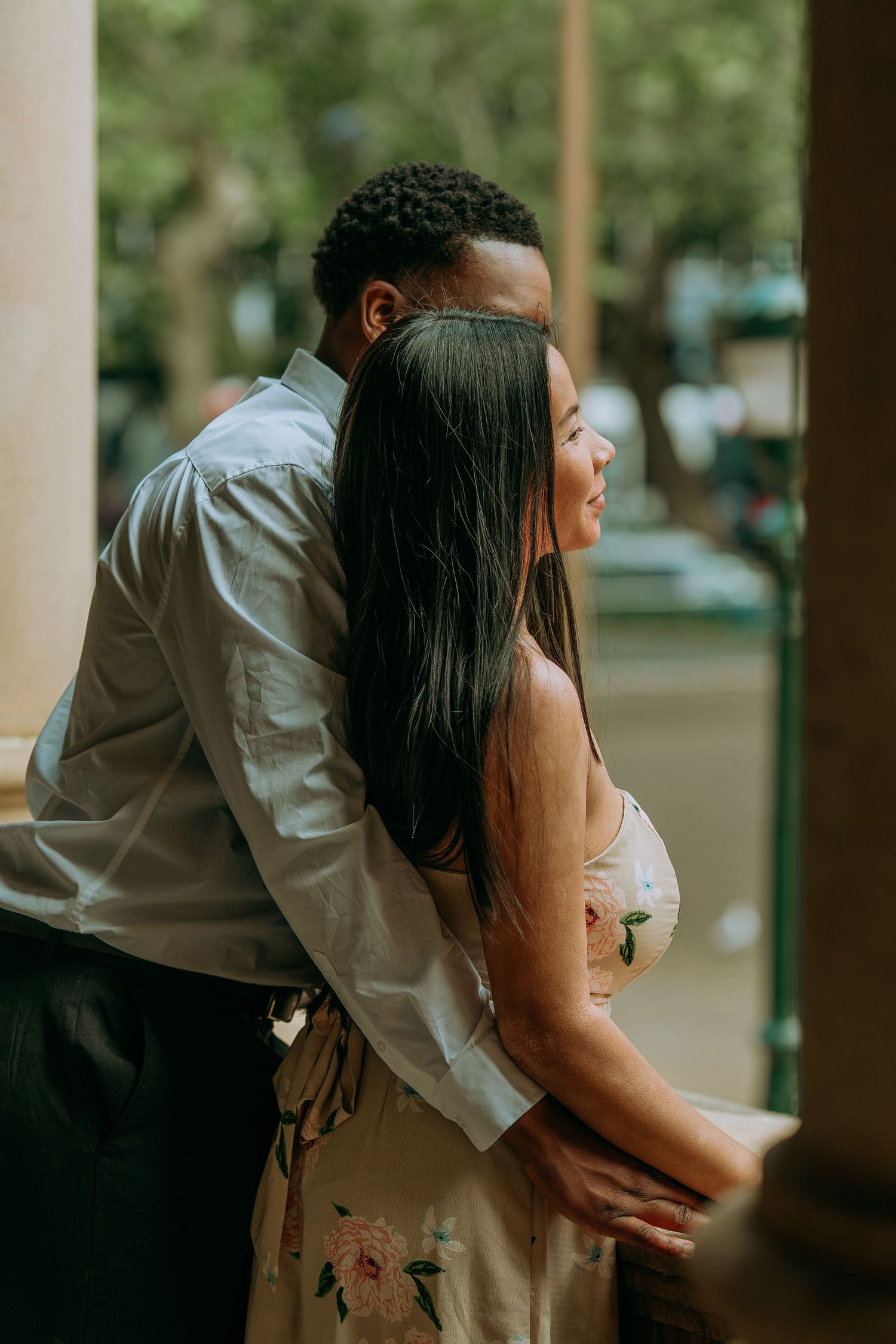 Couple embracing on a balcony overlooking a tree-lined street, woman smiling in floral dress.