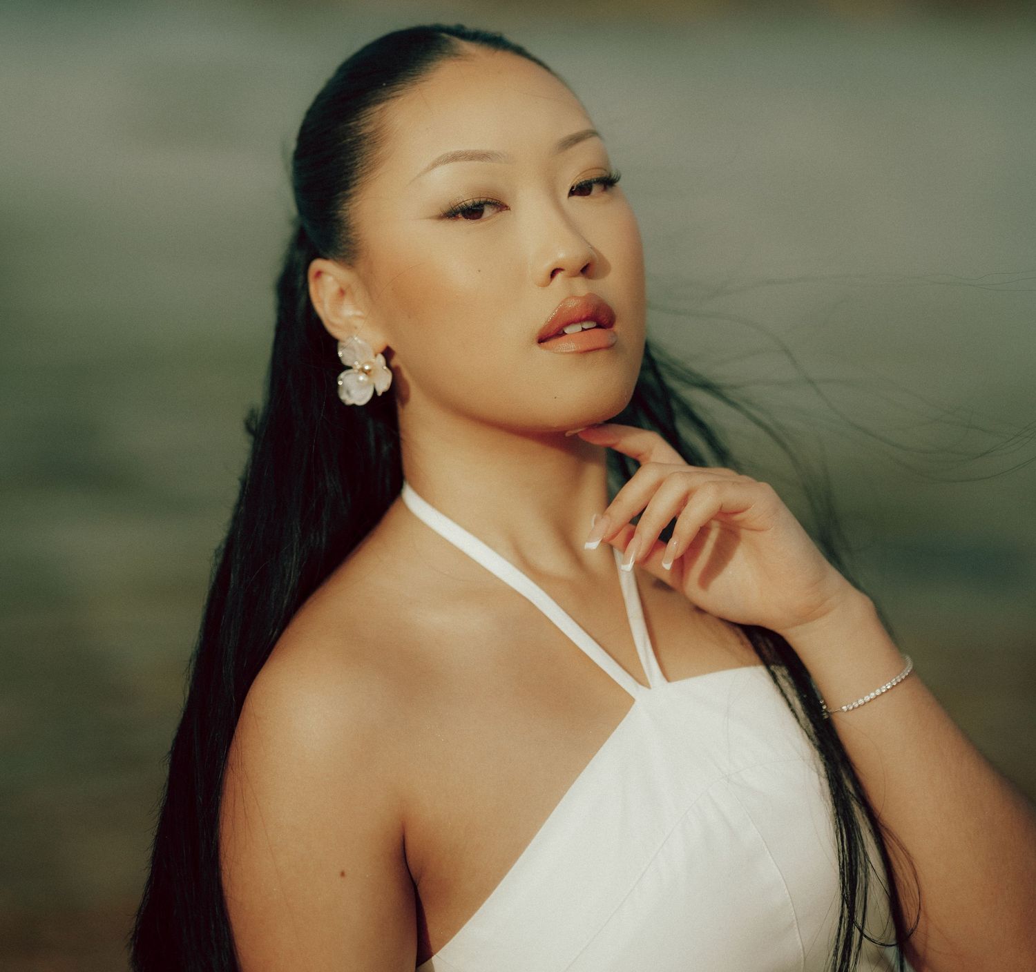 Portrait of Asian woman in white halter dress with flower earrings touching chin against blurred outdoor backdrop.