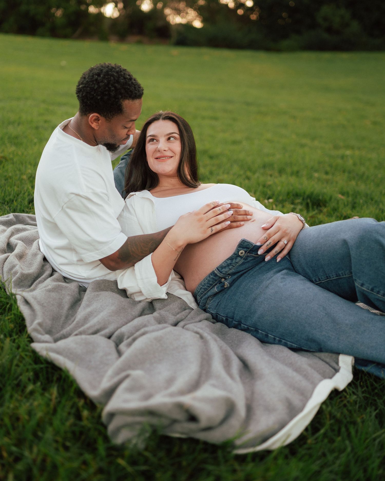Couple relaxing on a blanket in a green park during a casual outdoor maternity photo session.