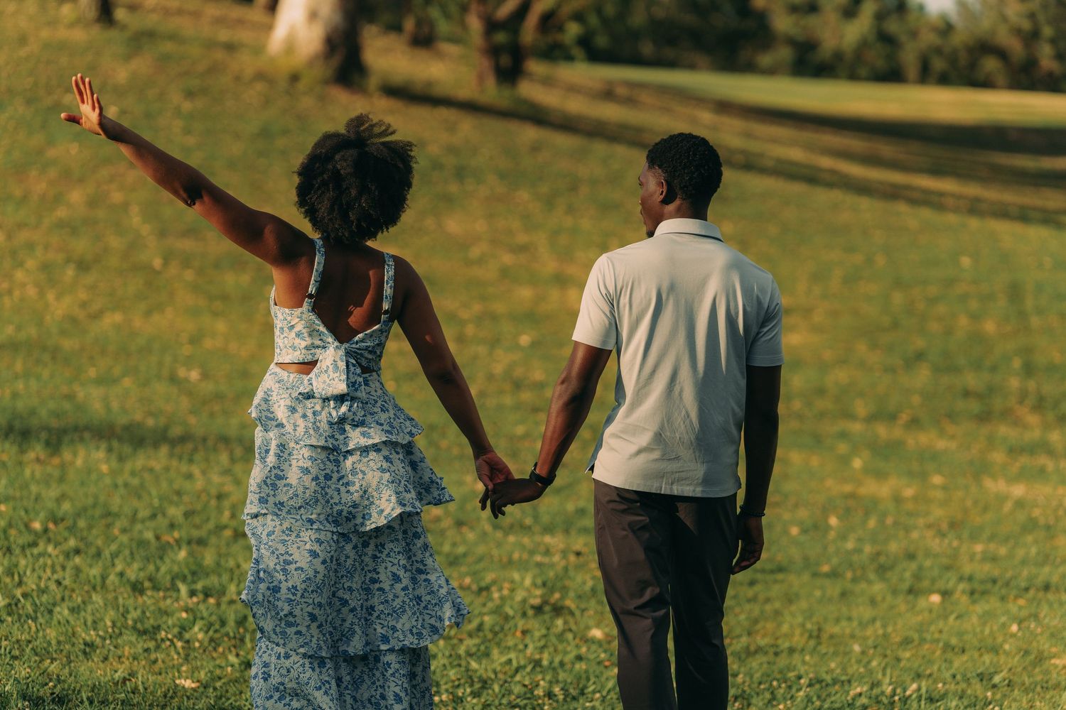 A couple walks hand in hand across a sunny green park, woman raising her arm joyfully.
