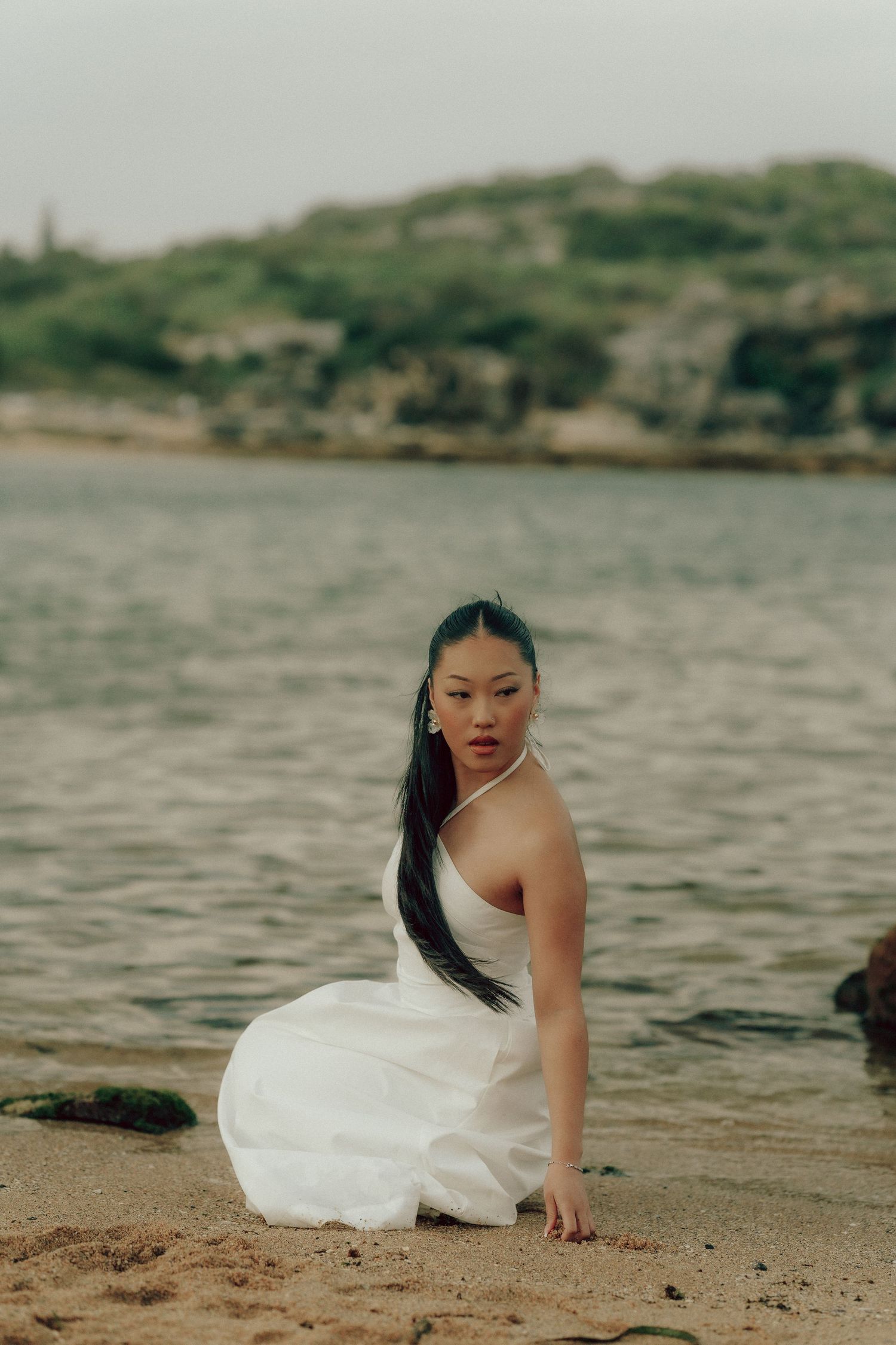 Young Asian woman in white halter dress sitting on sandy beach shore with rocky ocean backdrop.