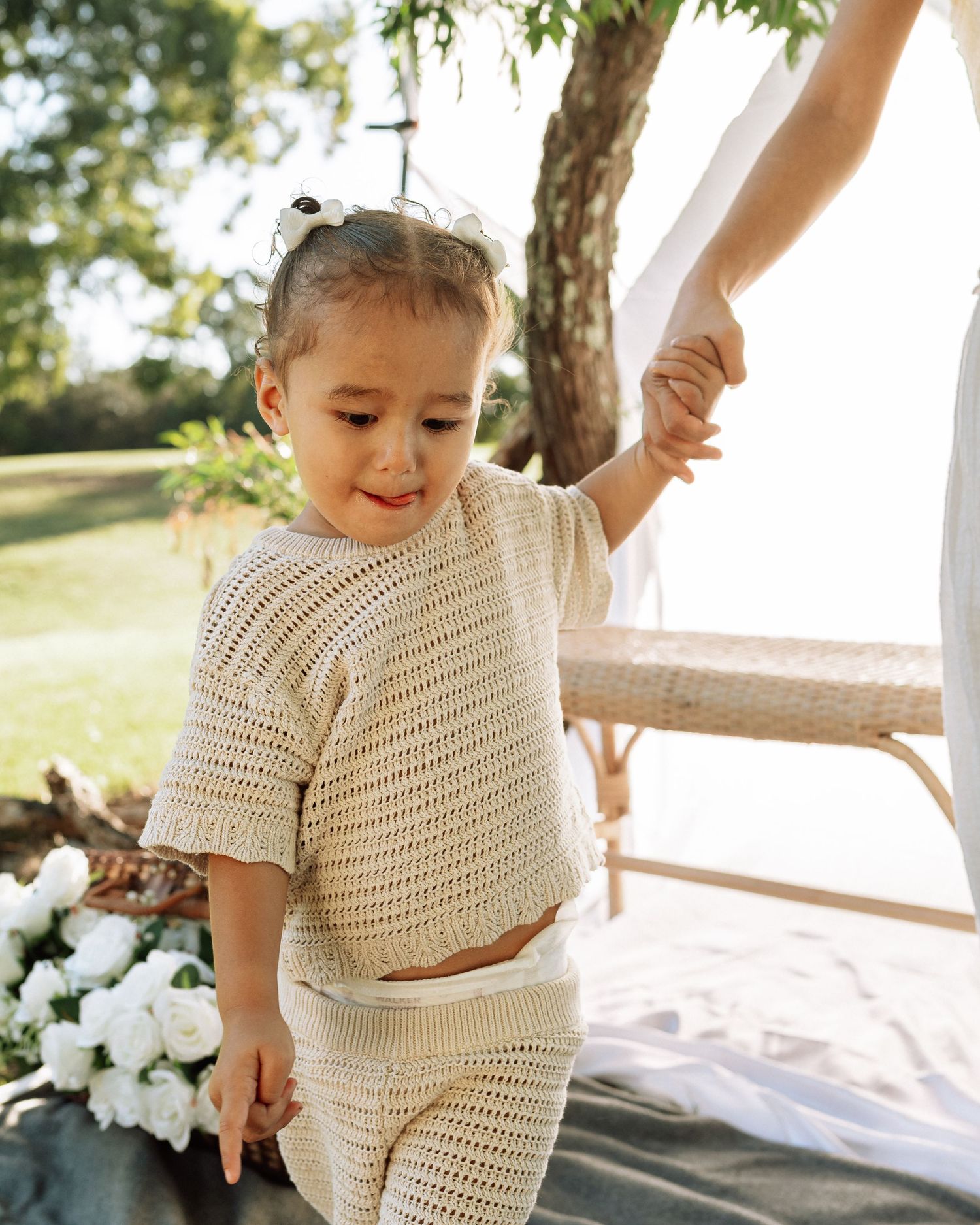 Adorable toddler in cream crochet knit outfit holding mothers hand near white floral arrangement in outdoor garden setting.