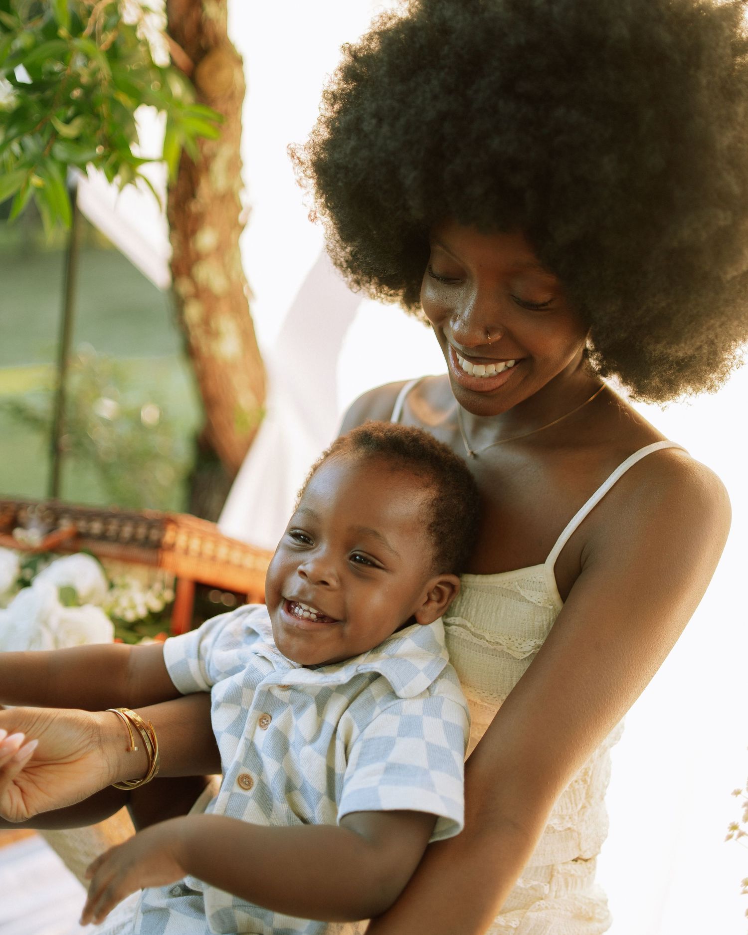 Joyful Black mother with natural afro holding her laughing baby boy in a sunny outdoor setting.