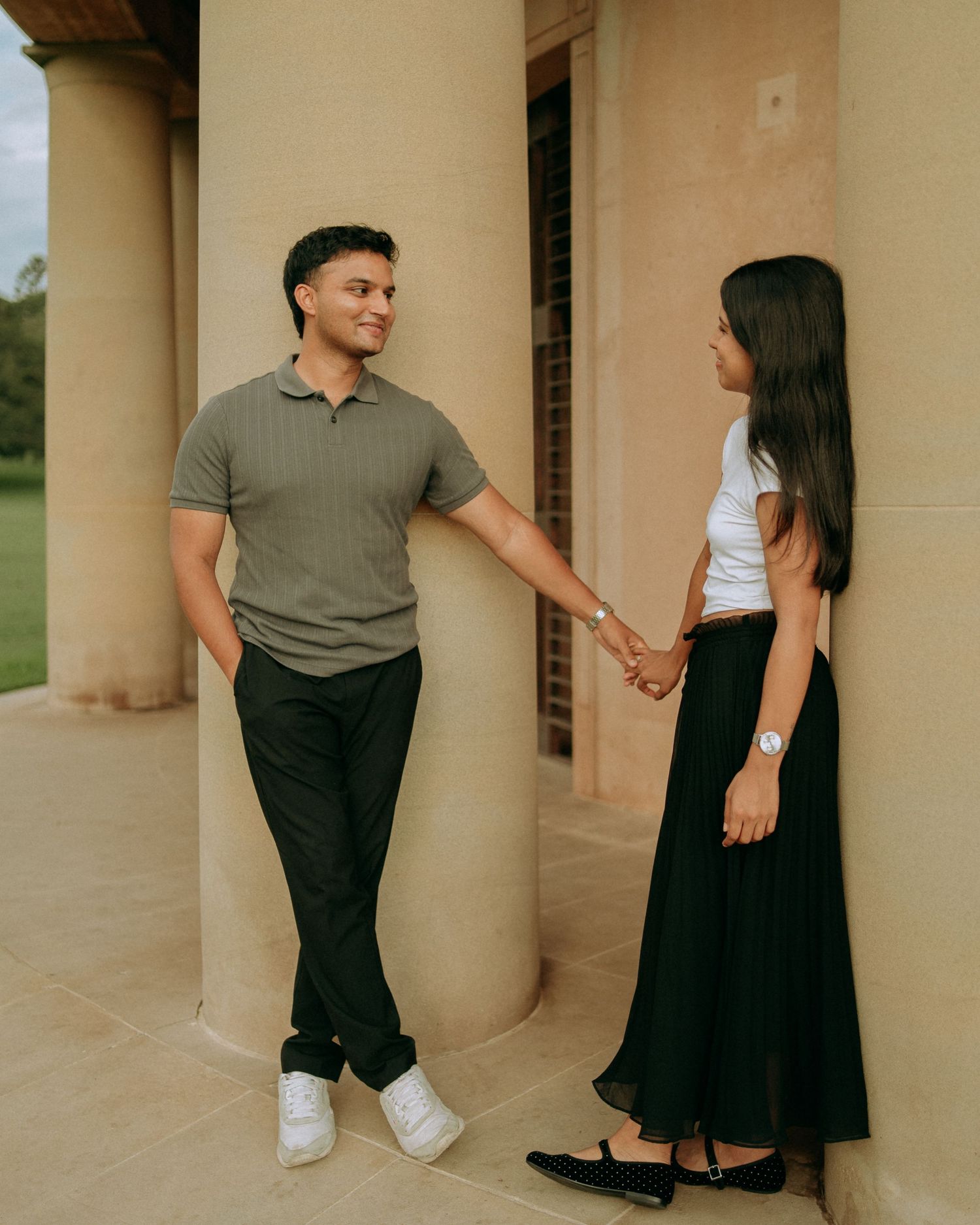 A young couple holds hands and smiles at each other while standing beside stone columns outdoors.