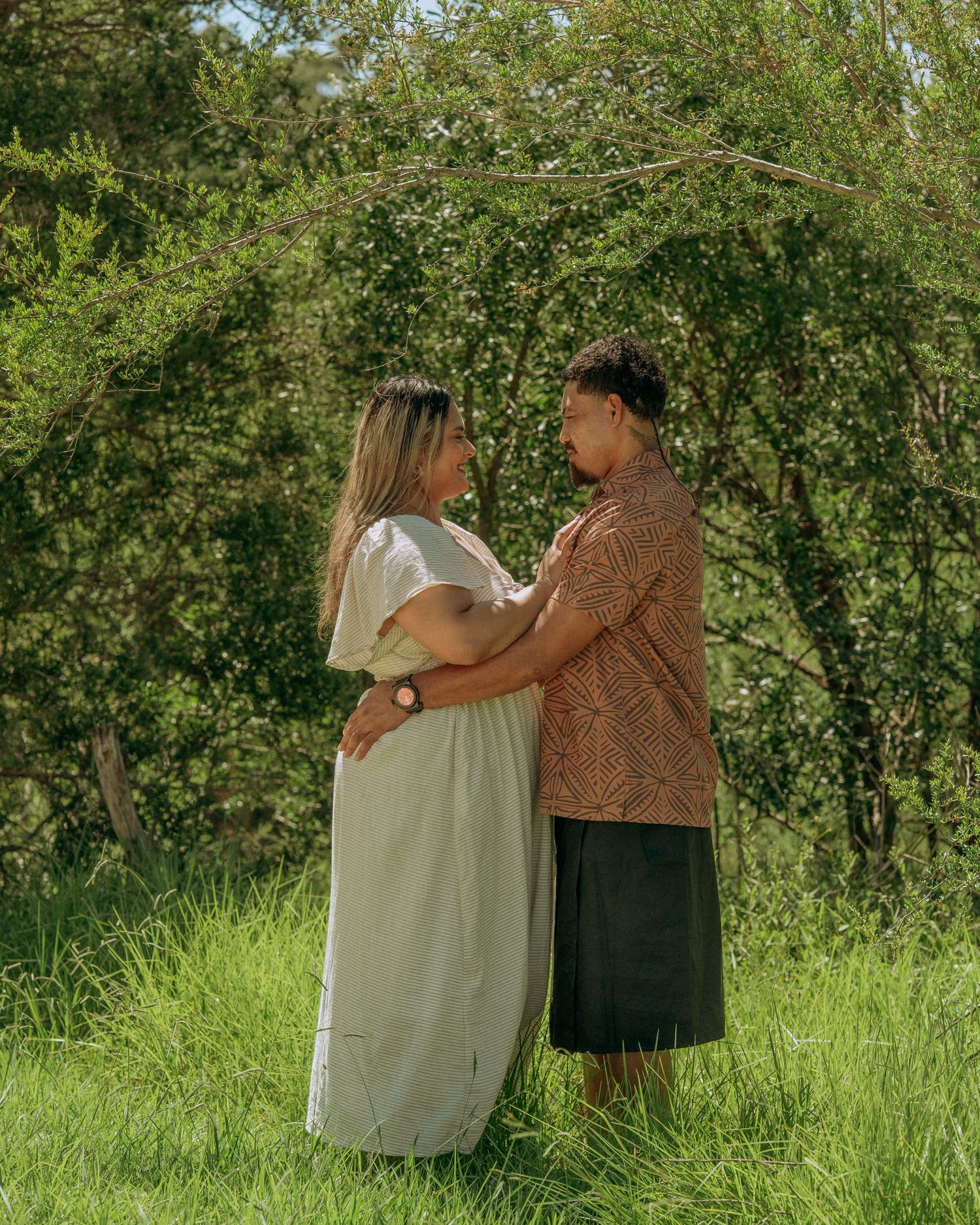 Couple embracing and gazing into each others eyes while standing in lush green bushland surroundings.
