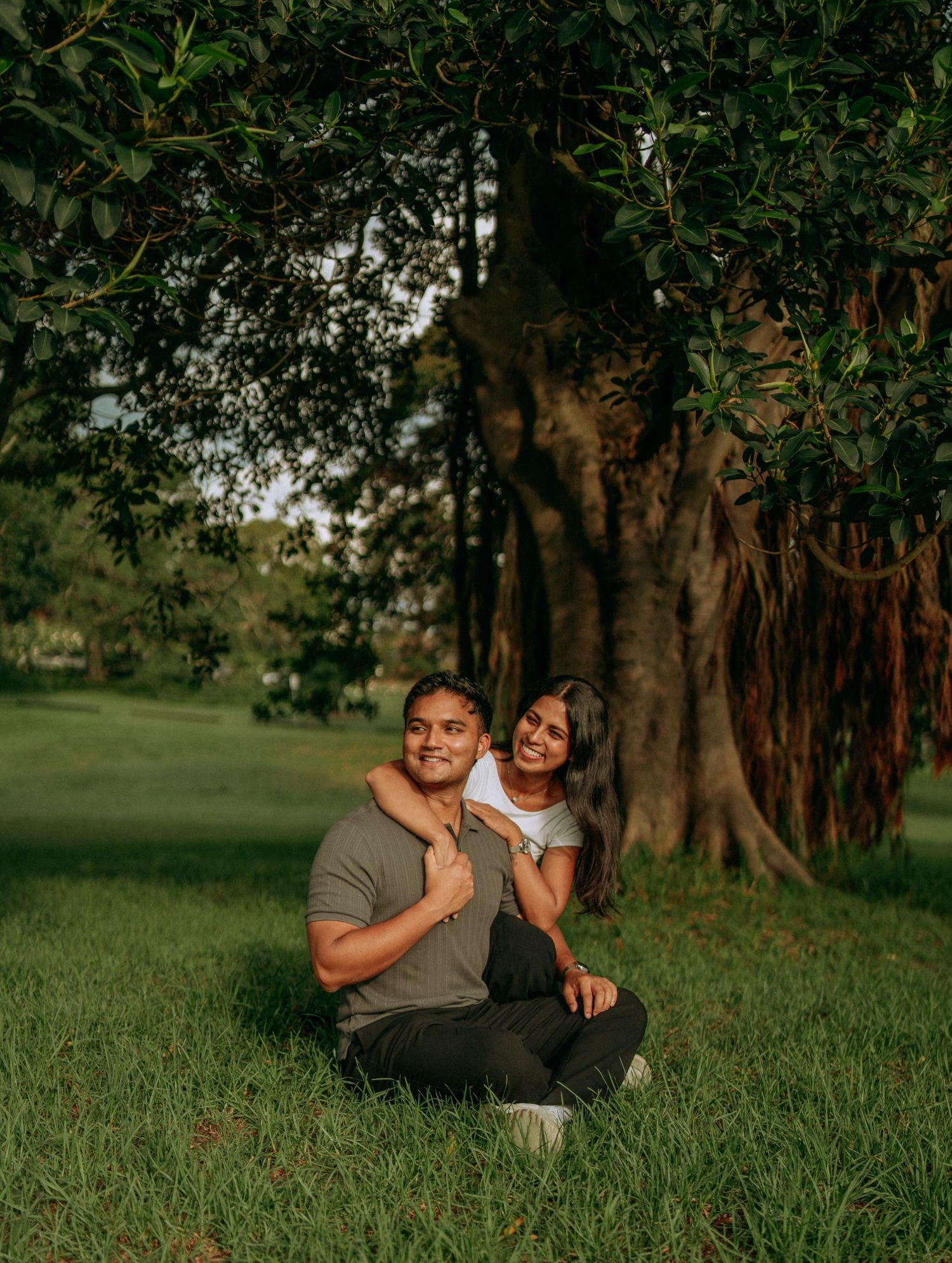 Couple sitting together smiling under a large tree in a green park.