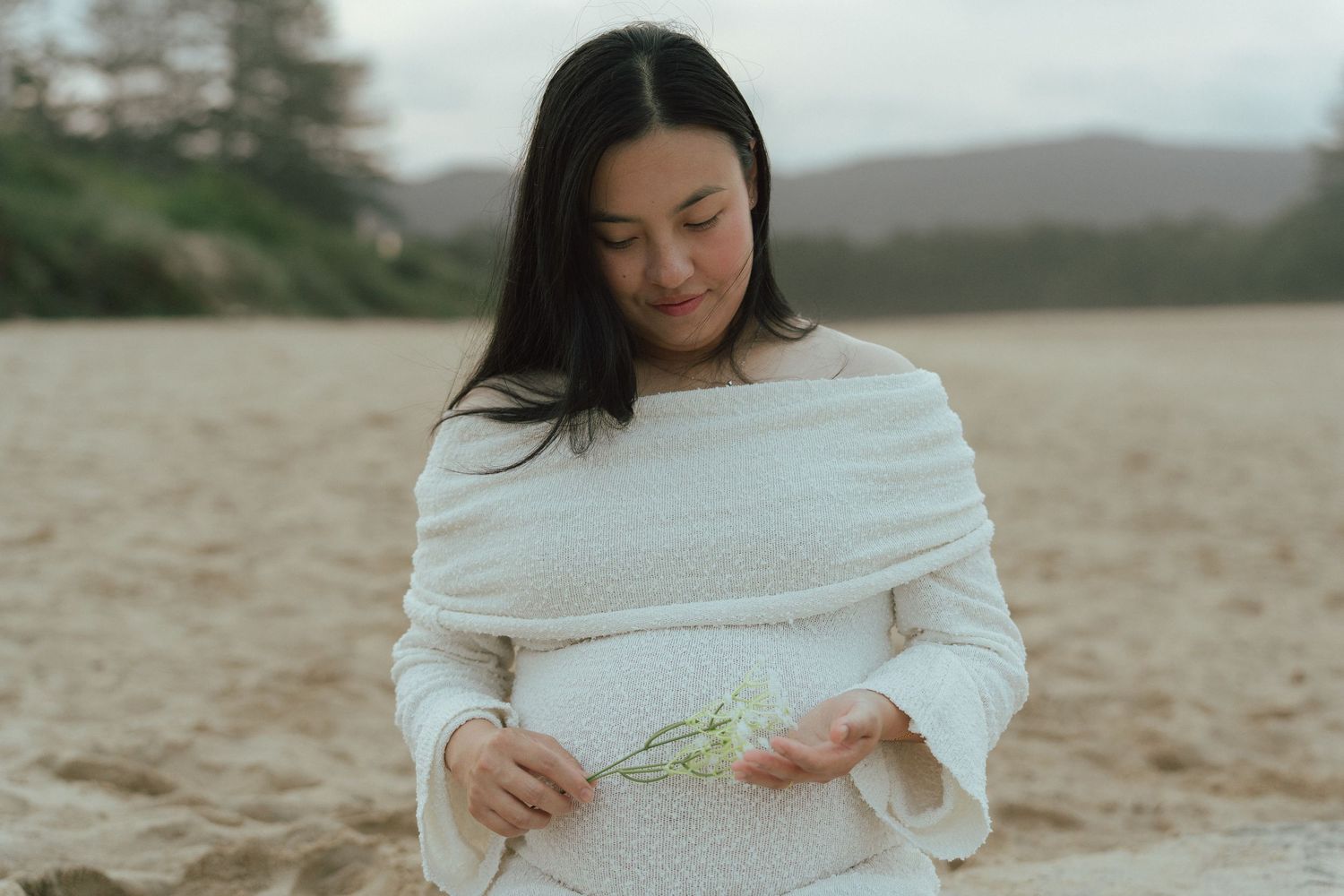 Pregnant woman in white off-shoulder knit dress holds a small flower on sandy beach during maternity session.