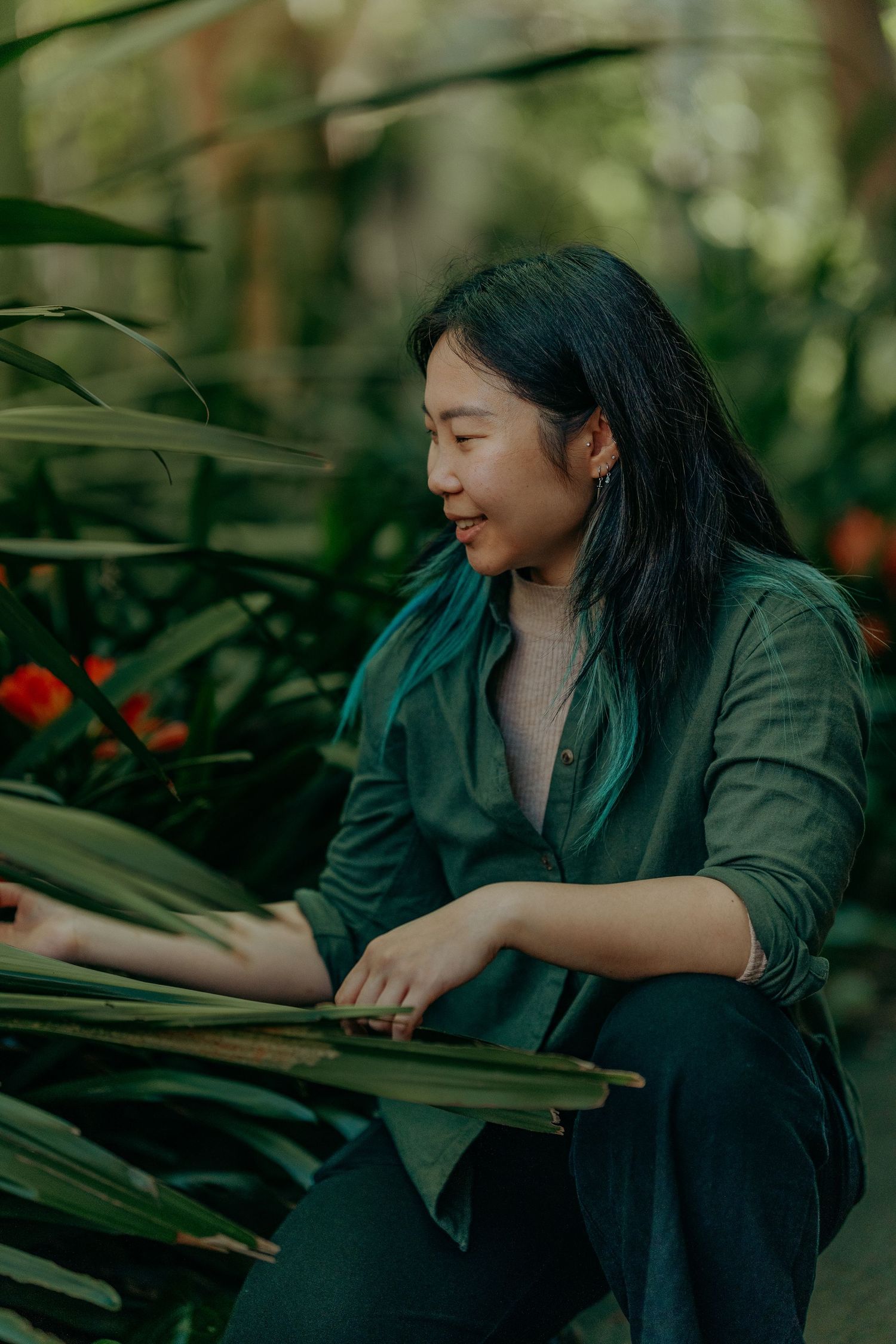 Smiling Asian woman in green shirt sitting among tropical plants in lush garden greenhouse.