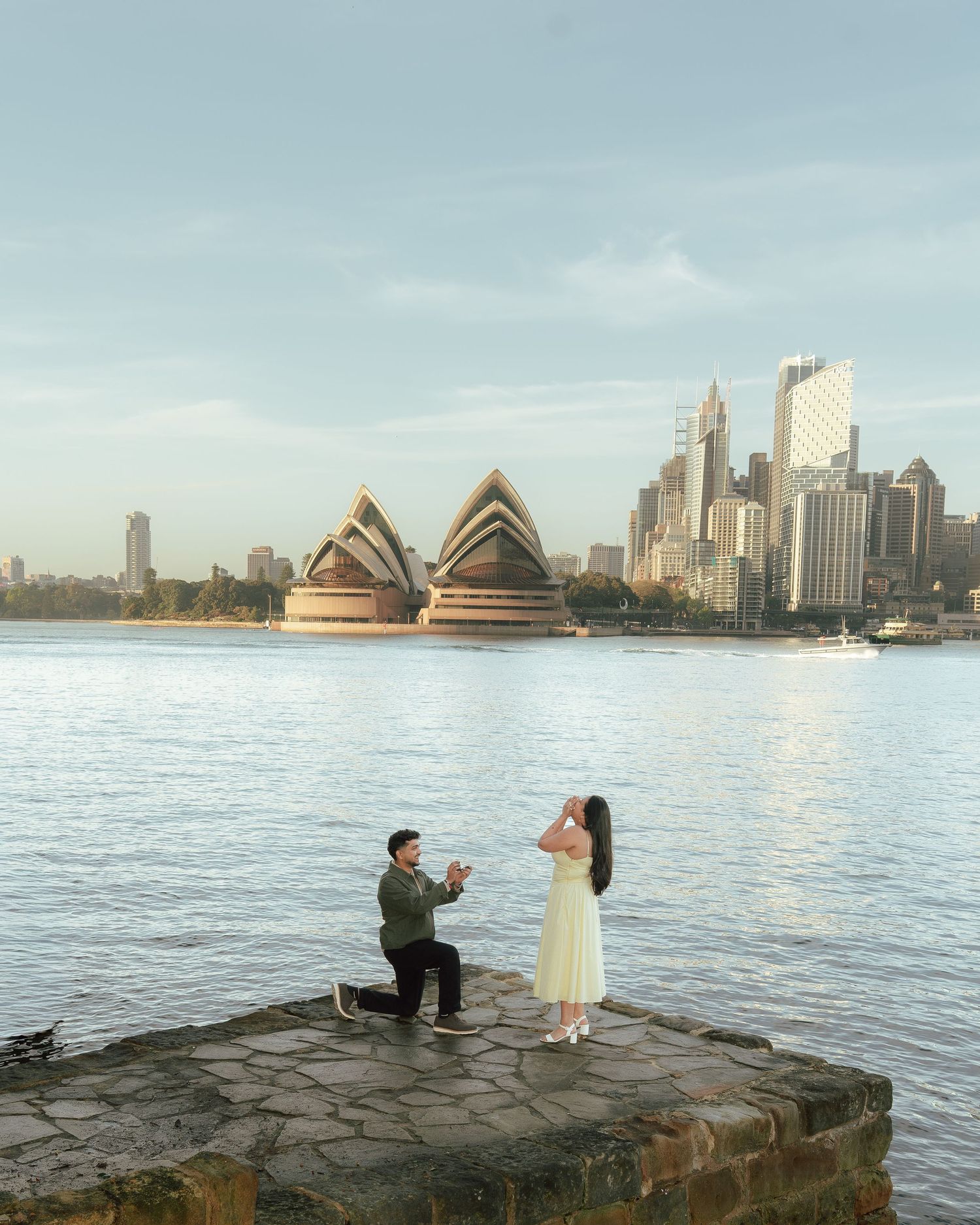 Man proposes to woman on rocky pier with Sydney Opera House and city skyline in background.