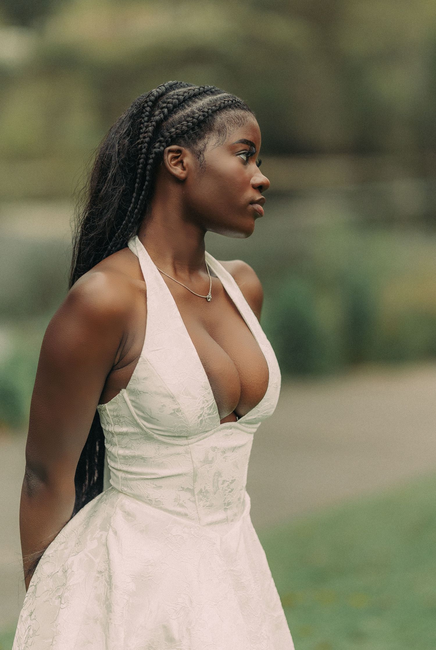 Young Black woman in white halter dress with long braids gazes sideways in an outdoor portrait.