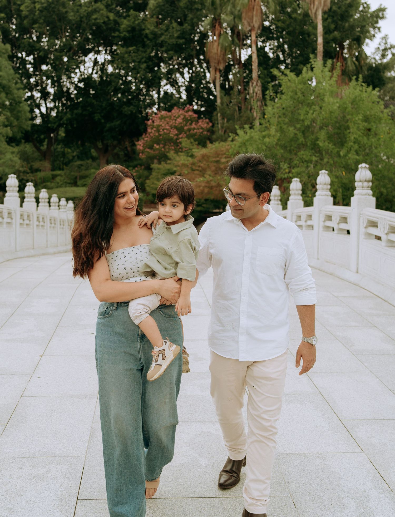 Young family with toddler walking on a white stone bridge surrounded by lush green trees.
