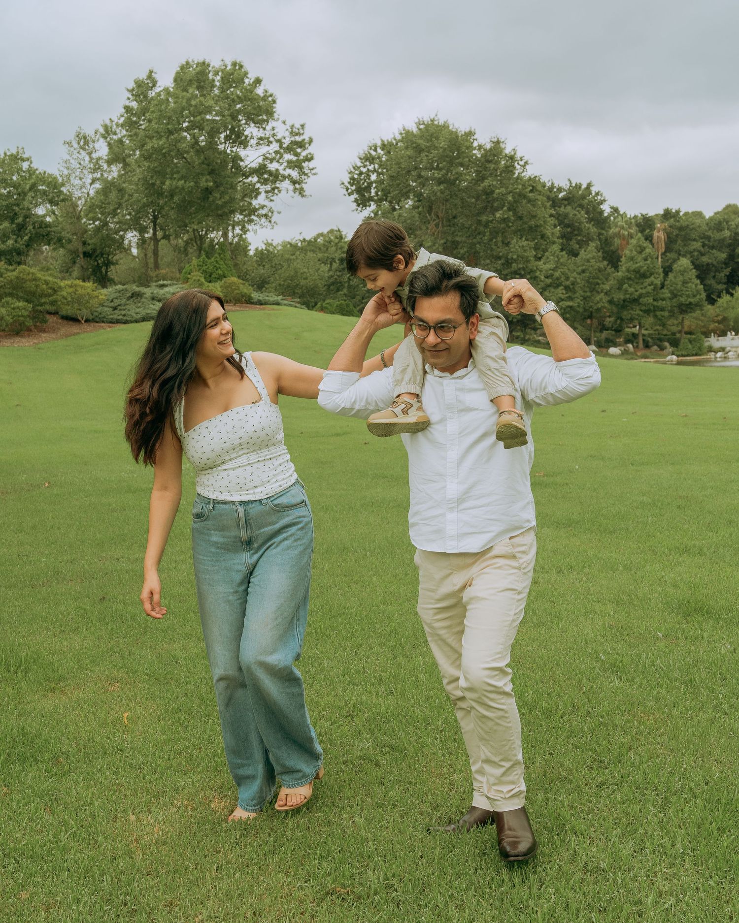 A happy family strolls across a green park lawn with their young child perched on the fathers shoulders.
