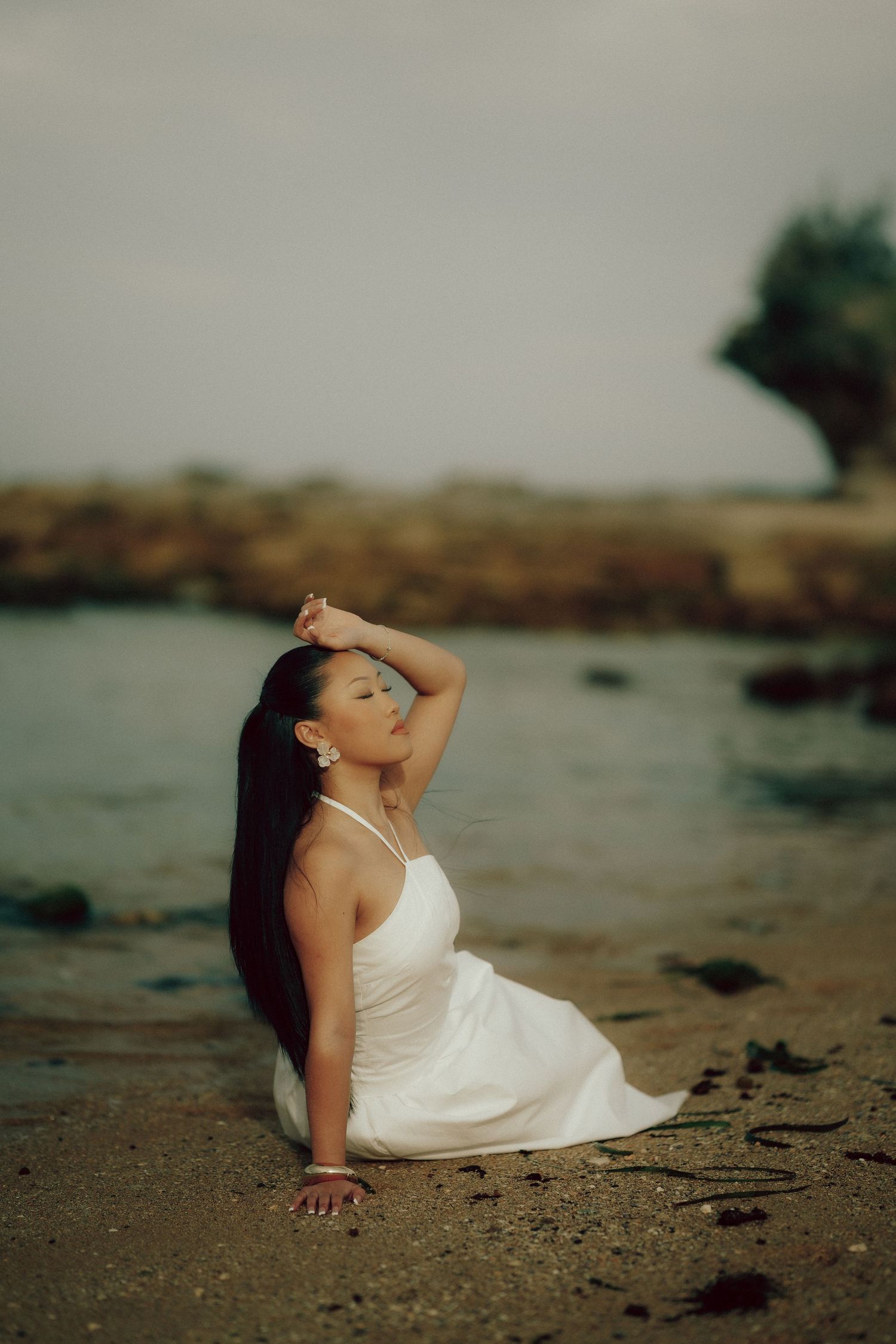 Woman in white halter dress sitting on wet sand by the shore with eyes closed and hand raised.