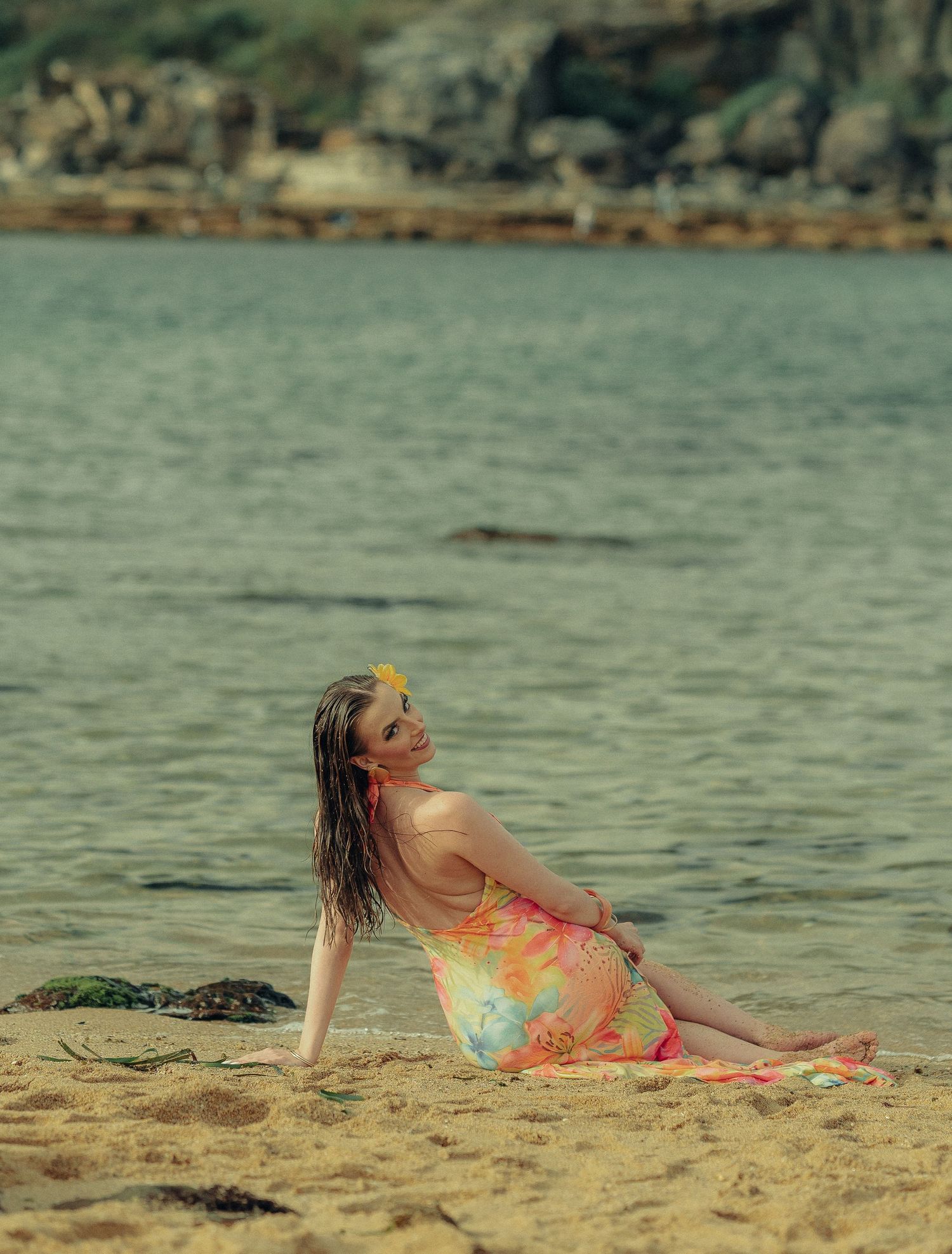 Woman in vibrant tropical dress sitting on sandy beach shore leaning back with ocean in background.