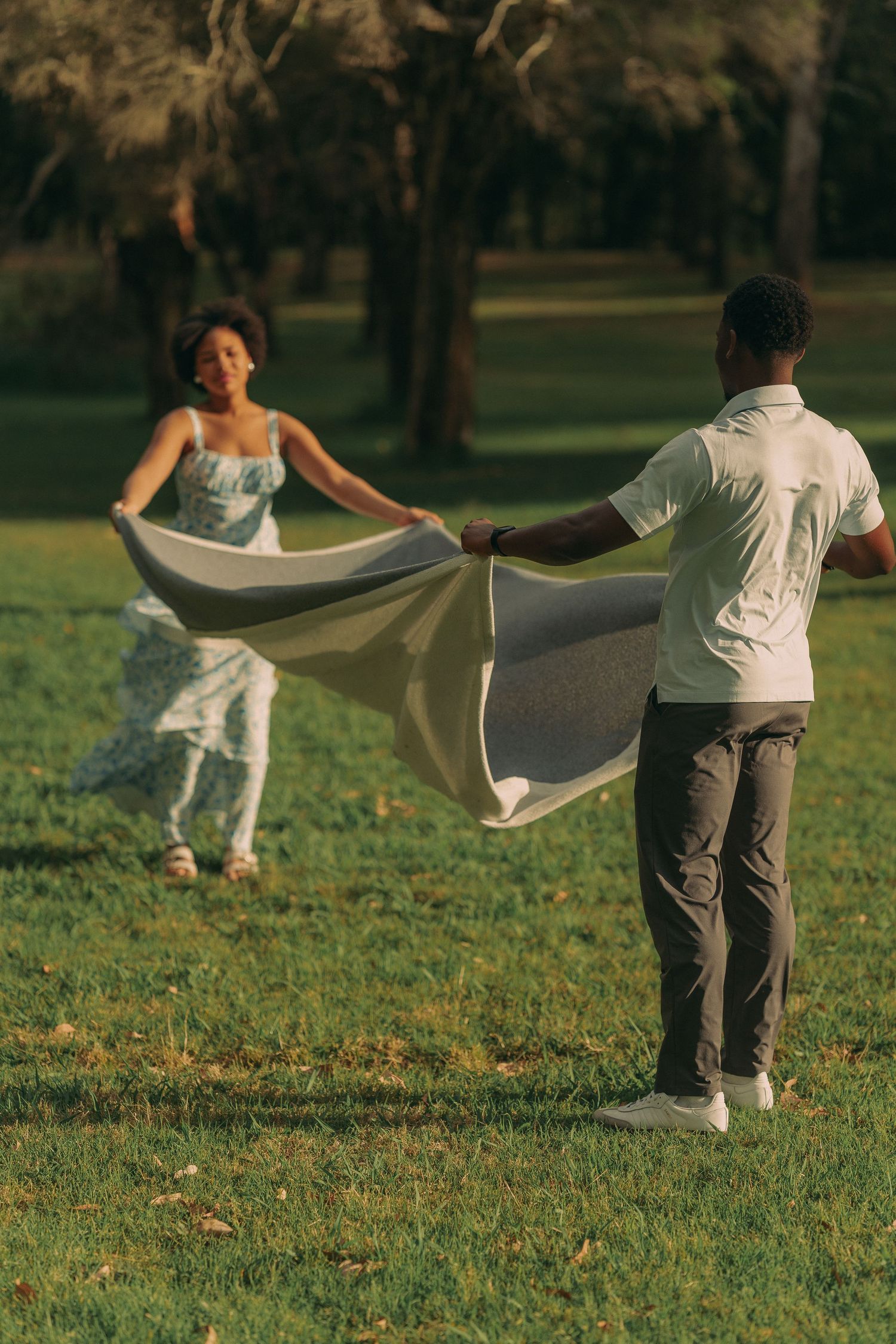 A couple playfully spreads a large blanket across a green park lawn in warm golden afternoon light.