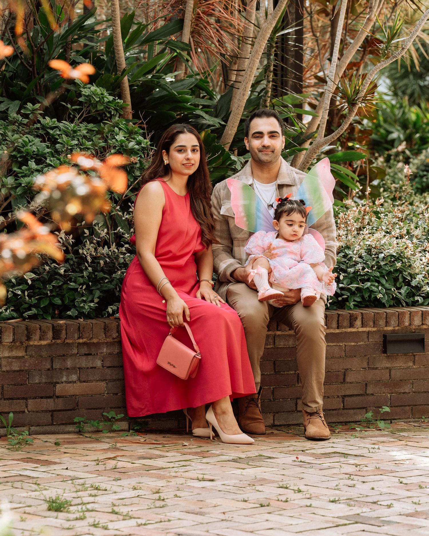 A stylish family poses together on a brick ledge surrounded by lush garden greenery, mother in a red dress.