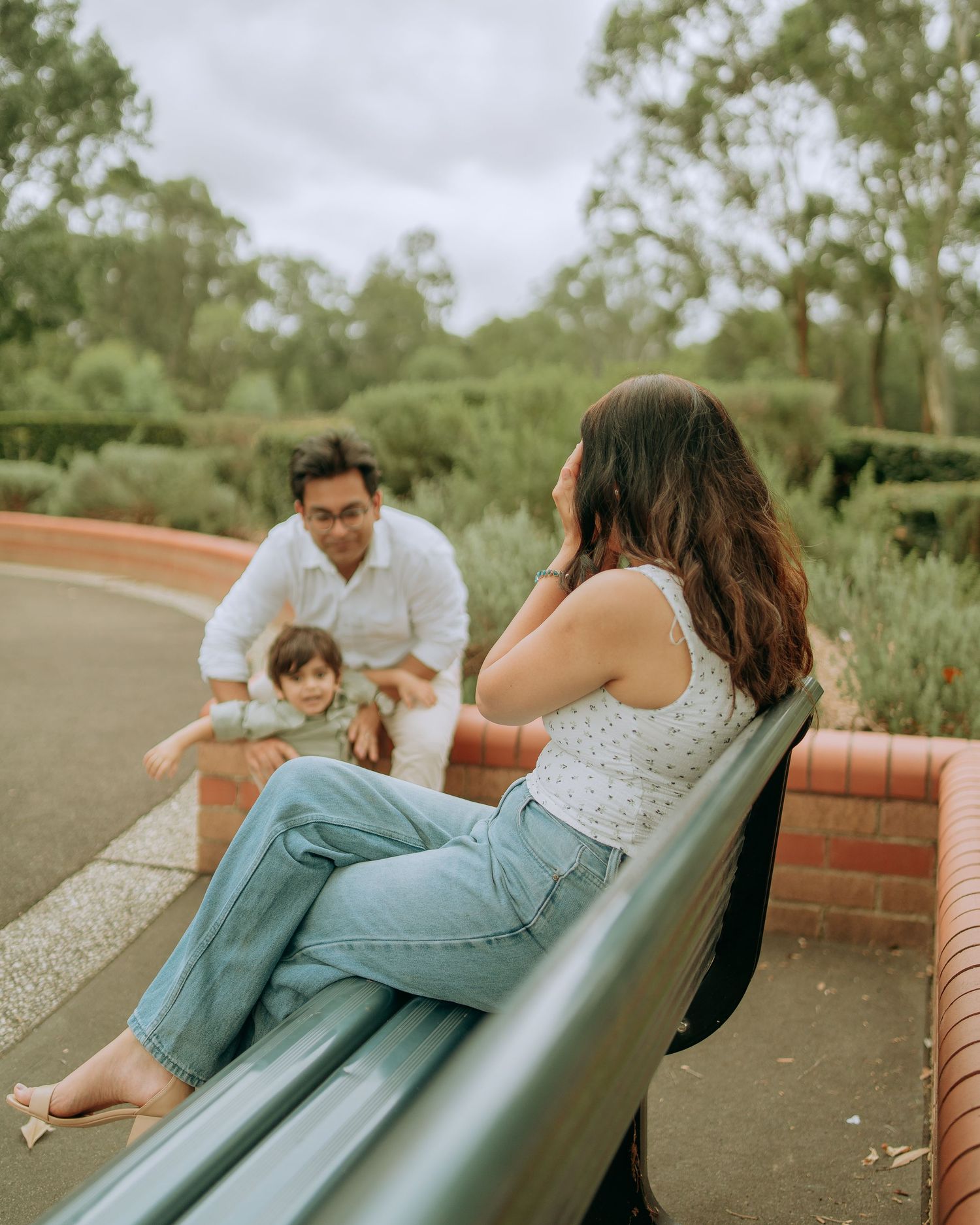 Father playing with toddler in park while mother sits on bench watching in a green outdoor setting.
