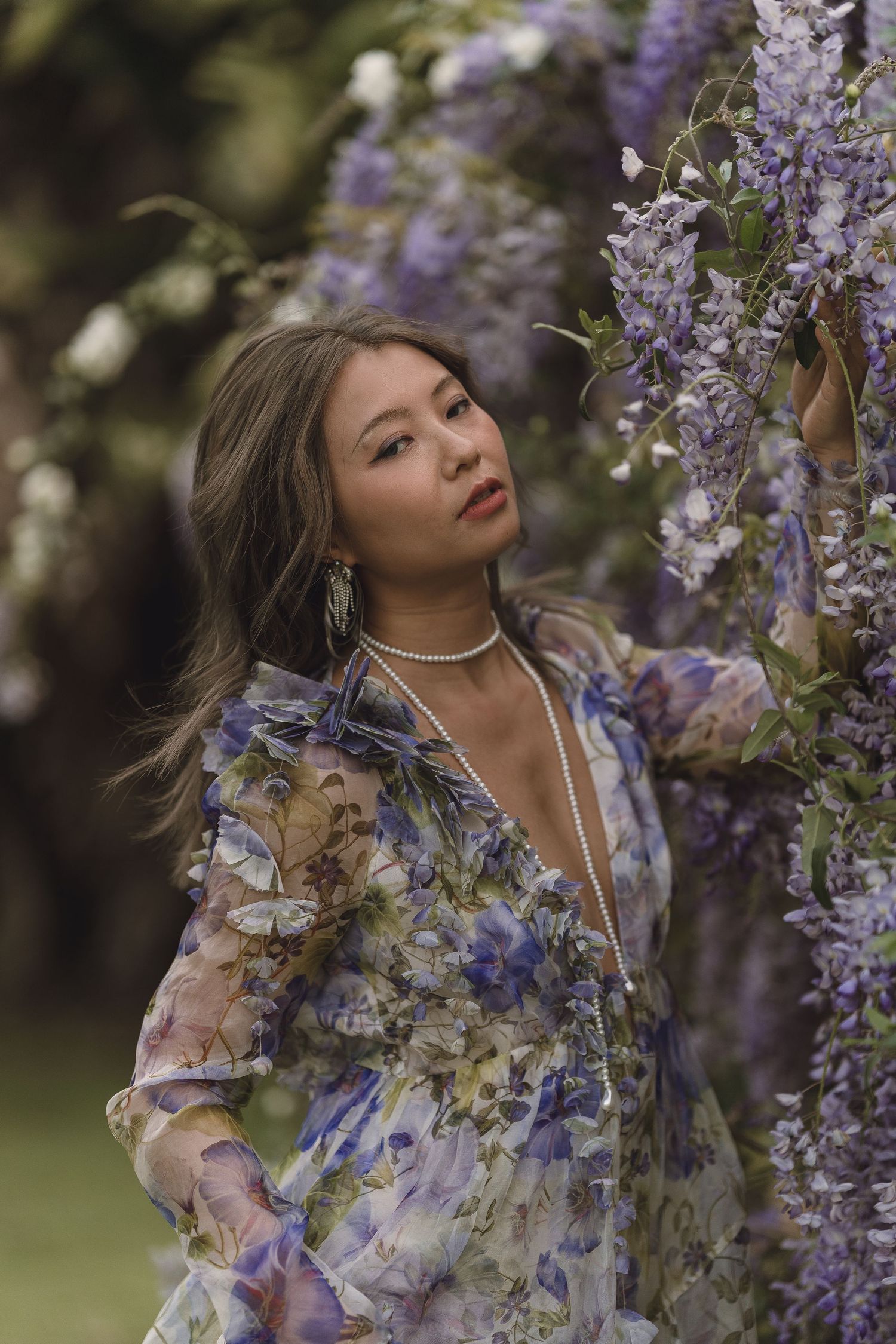 Asian woman in floral dress gazing up at cascading purple wisteria flowers in garden.