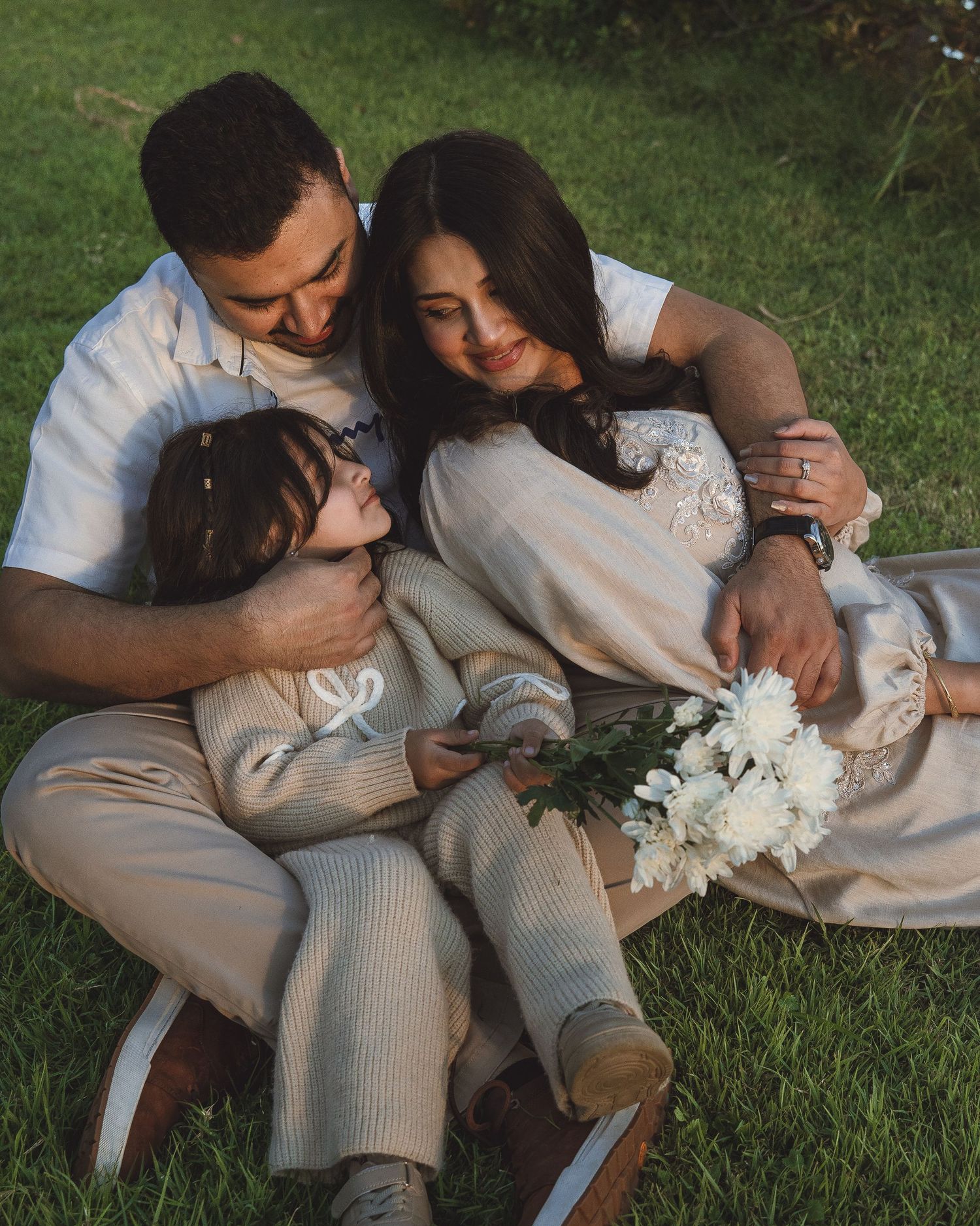 A loving family sits together on green grass, parents embracing their young child who holds a bouquet of white flowers.