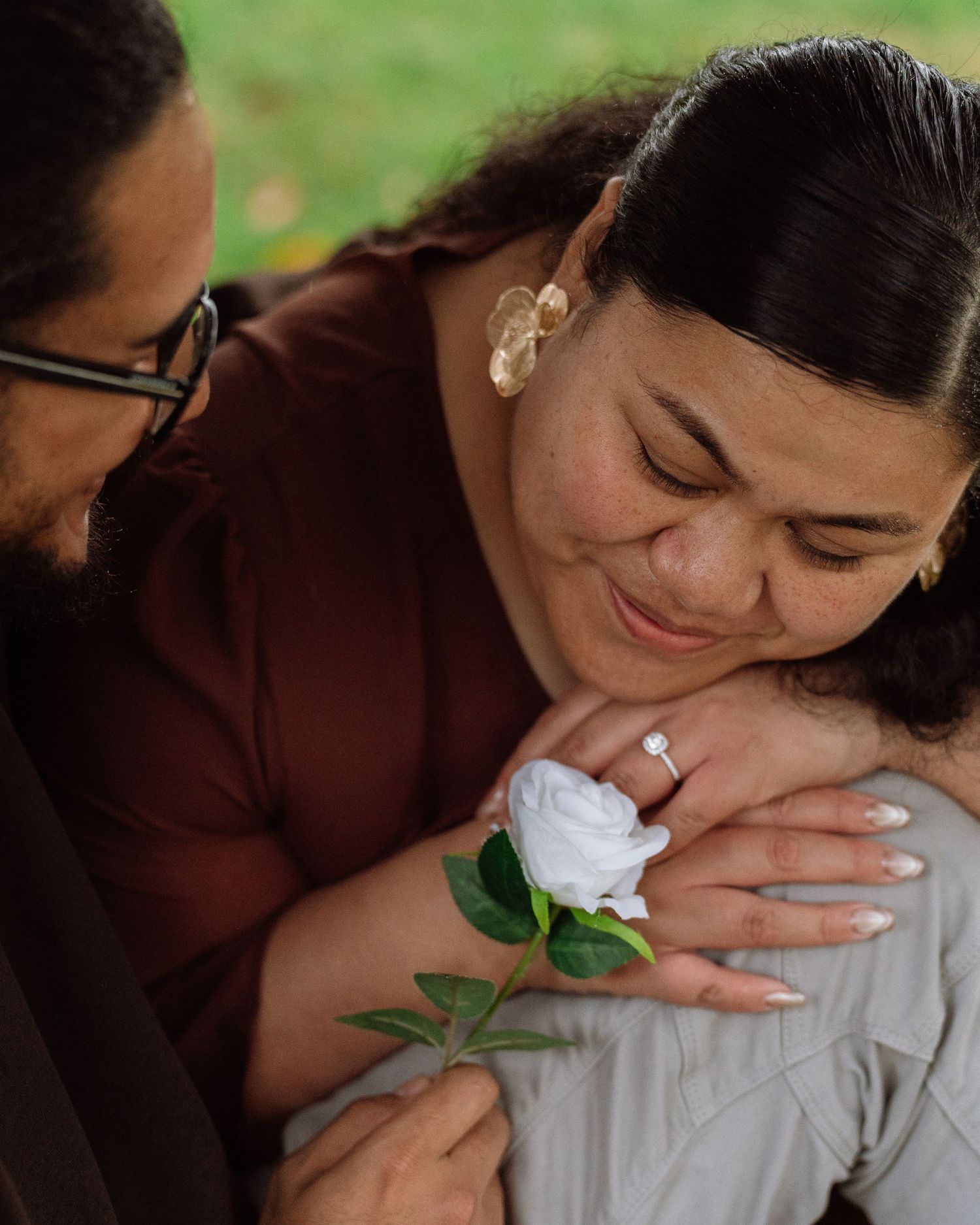 A man embraces a smiling woman from behind, presenting a white rose with an engagement ring visible.