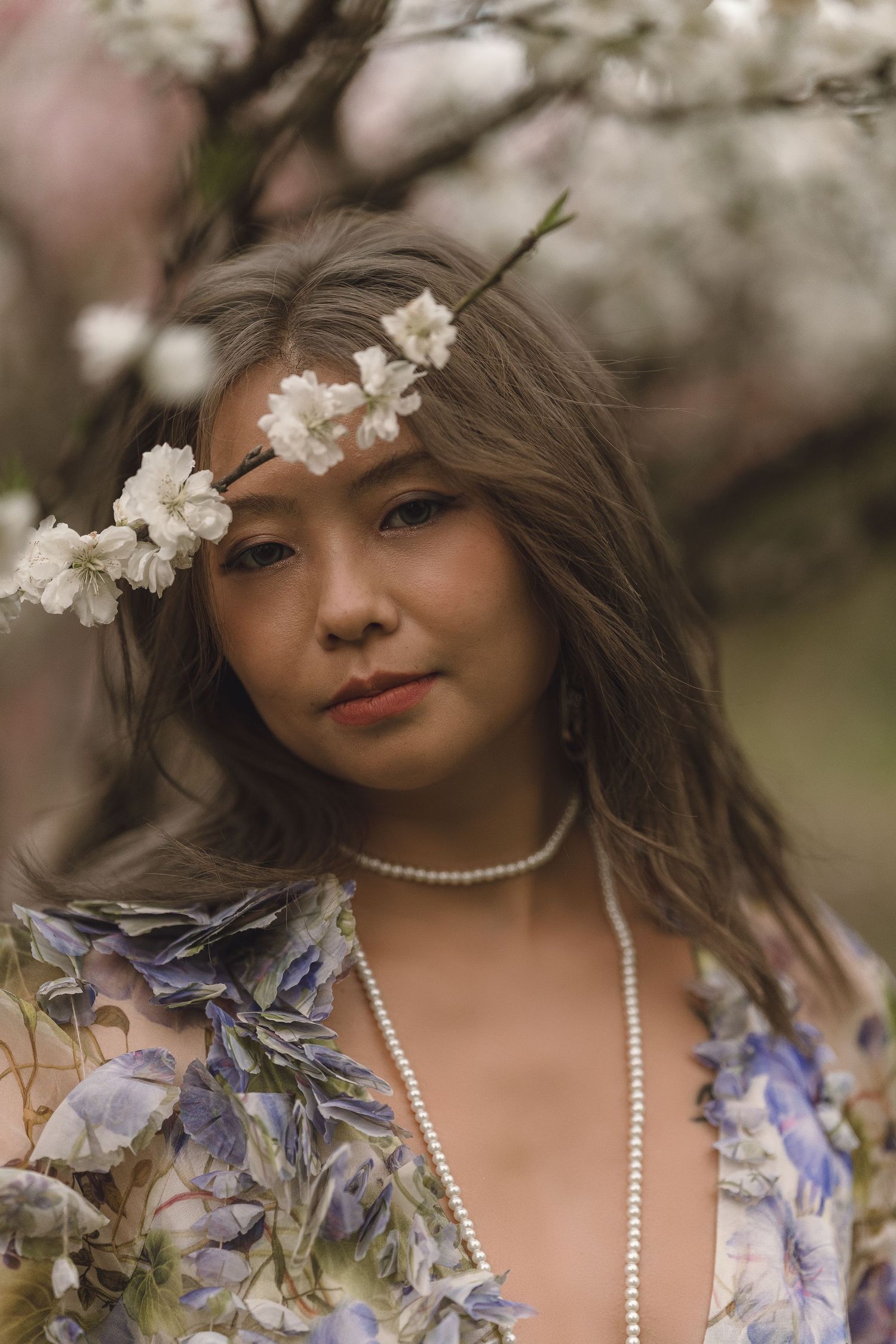 Asian woman with white cherry blossoms in hair wearing floral dress and pearl necklace in spring garden.