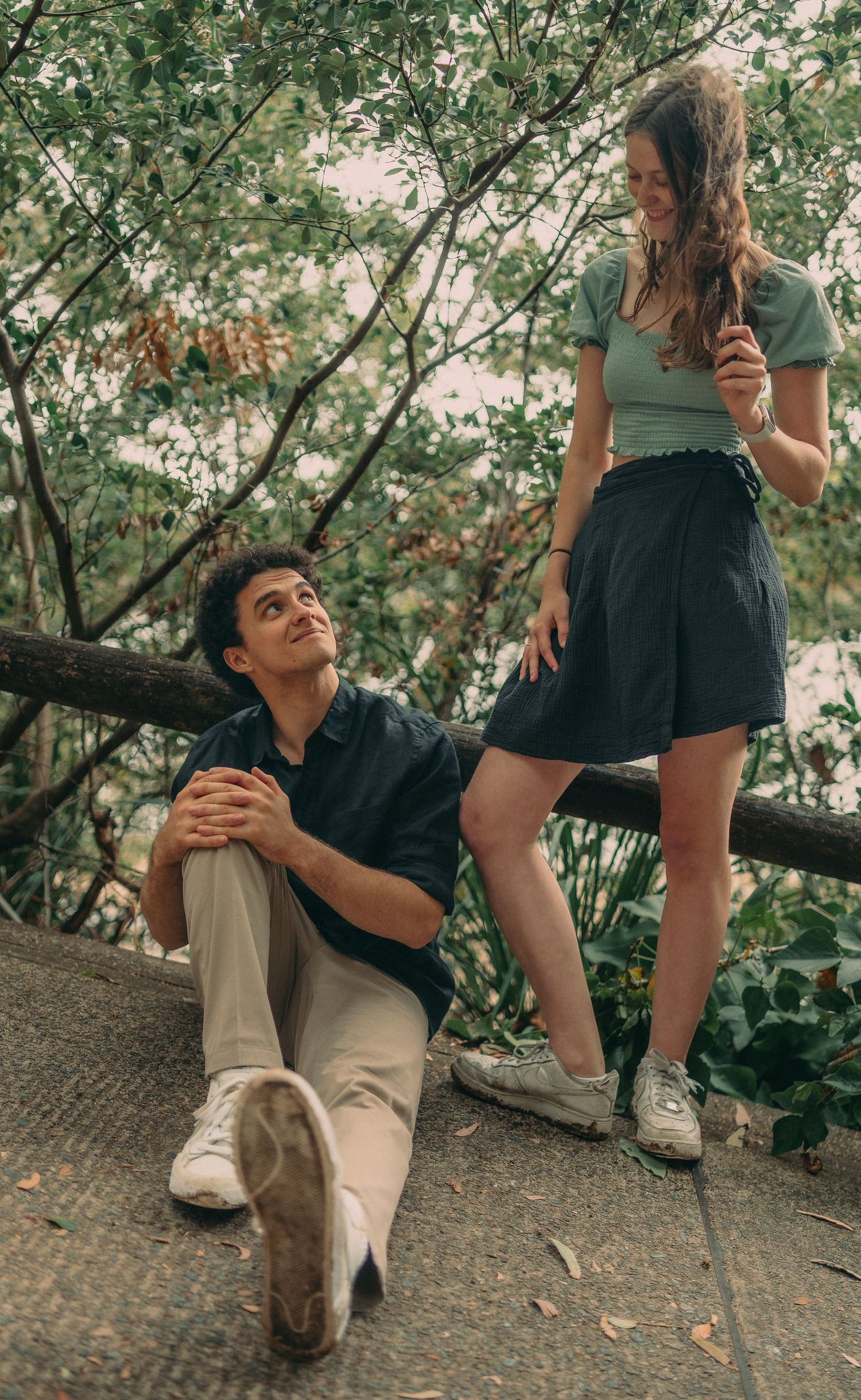 Couple in a lush outdoor setting, man sitting on ground looking up at smiling woman standing above him.