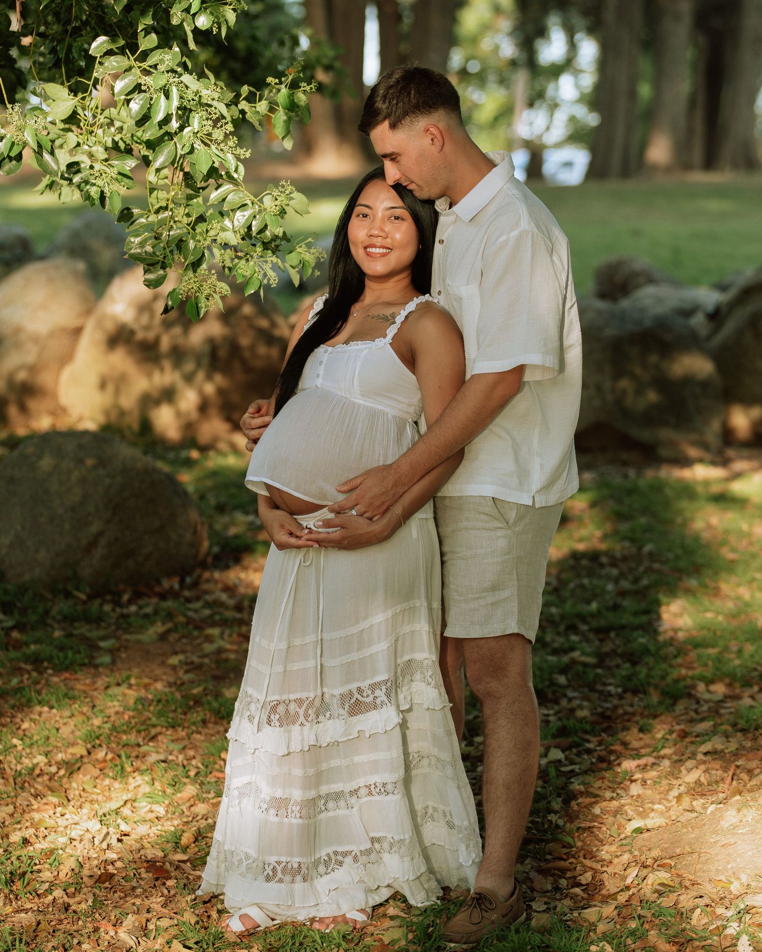 Pregnant couple in white outfits embracing outdoors in a sunny park during a maternity photo session.