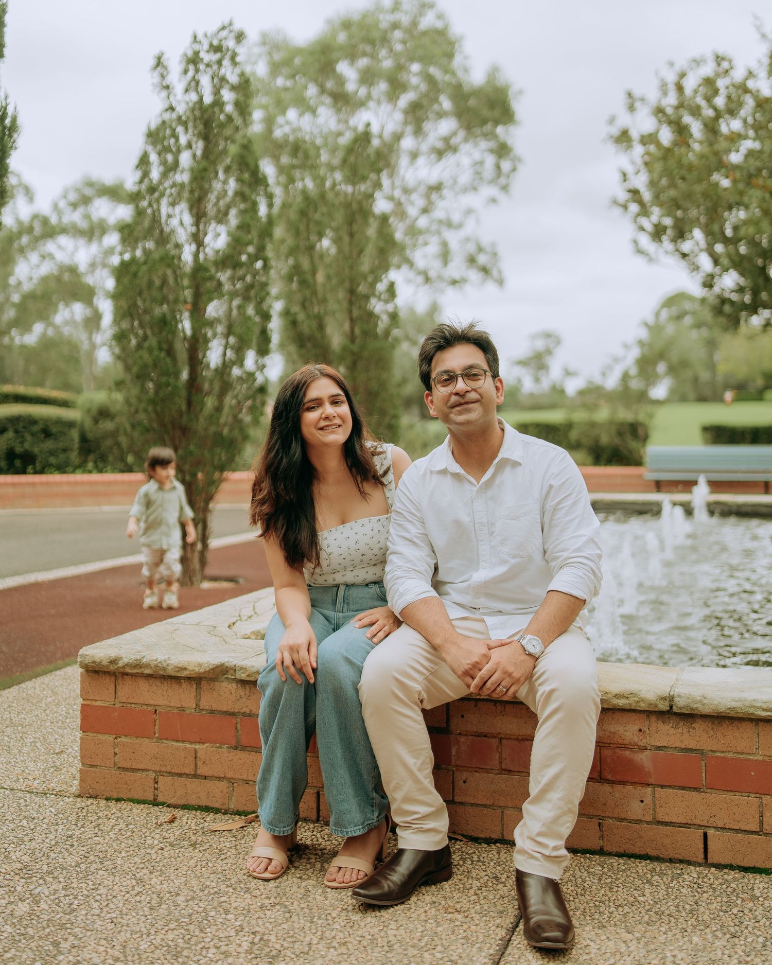 Couple sitting together on brick wall near fountain in park, with toddler playing in the background.