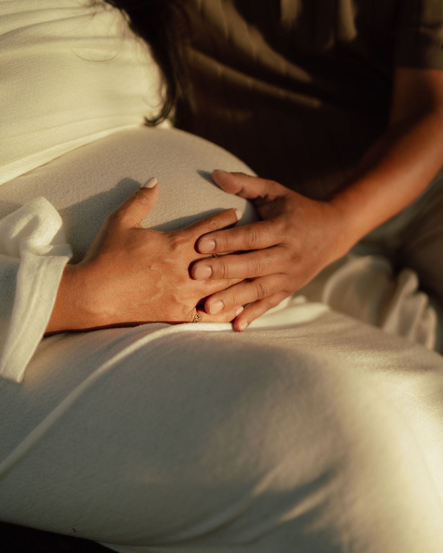 Pregnant woman in white dress with hands gently resting on baby bump in warm golden sunlight.