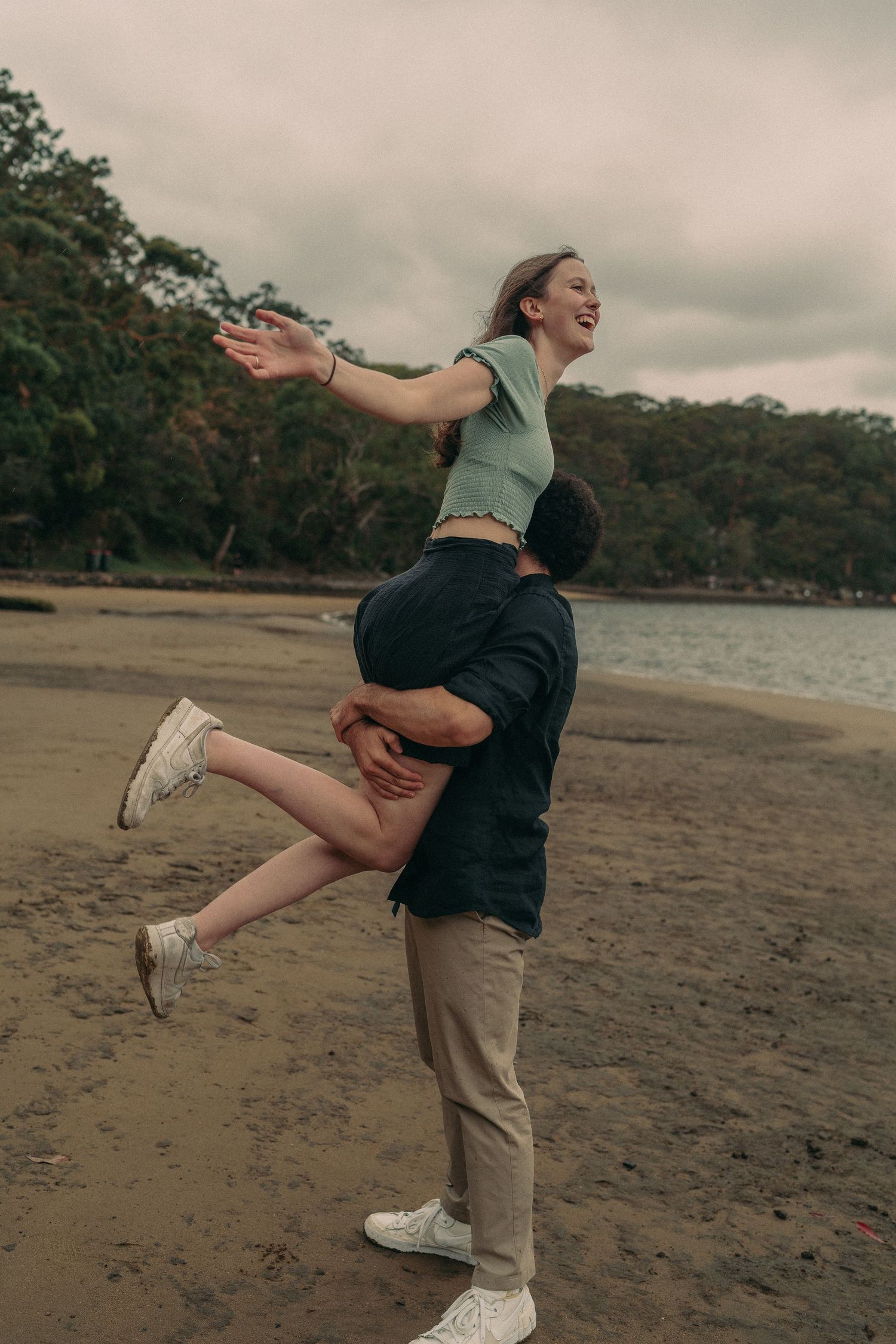 A joyful couple on a beach, man lifting woman who spreads her arms wide, smiling against cloudy sky.