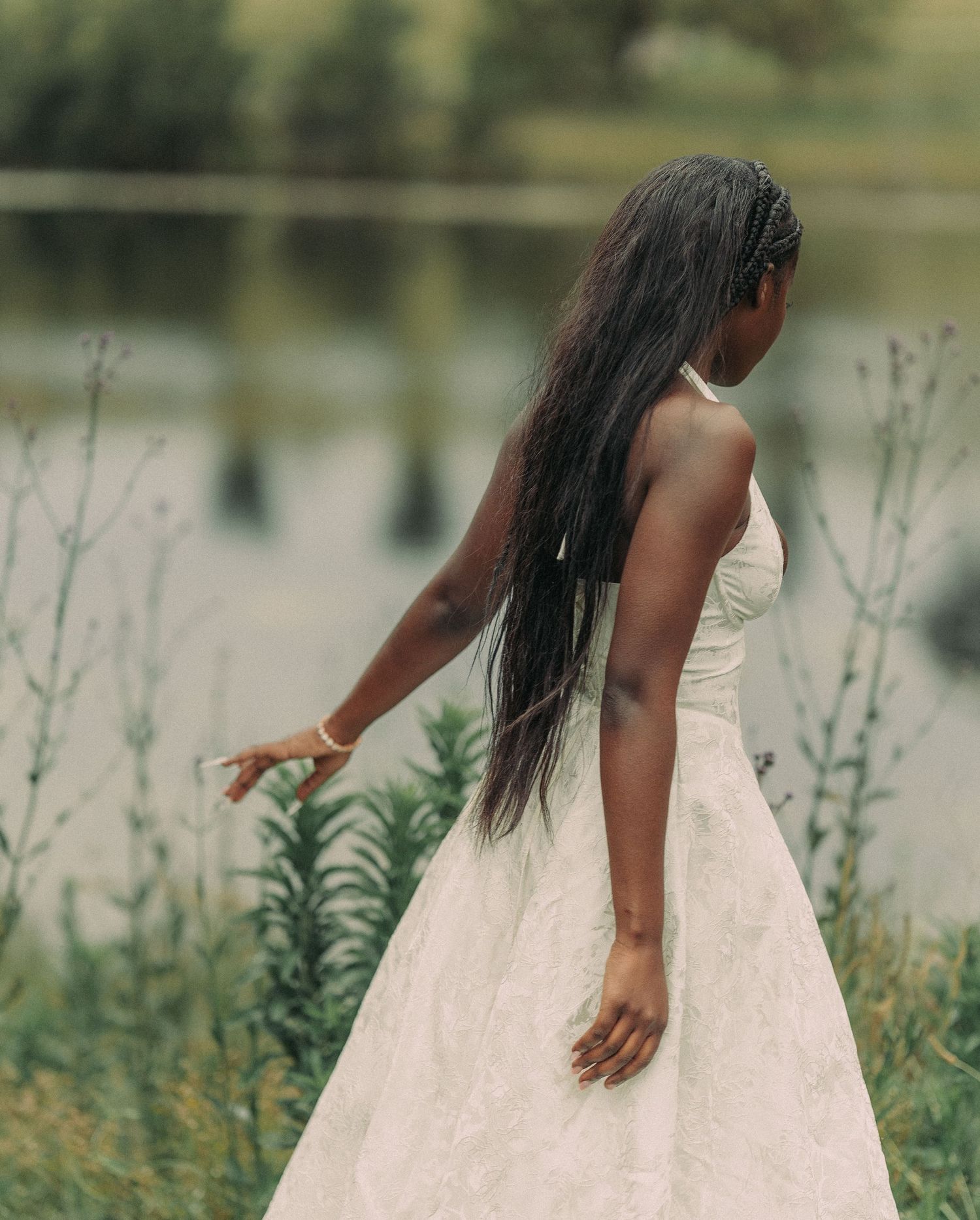 A young Black woman in a white halter dress stands among tall green plants beside a calm reflective lake.