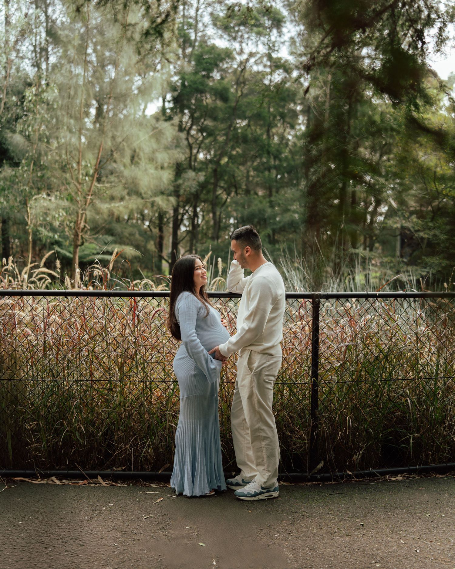 Couple holding hands in a park, woman in light blue maternity dress, man in cream outfit, surrounded by lush greenery.