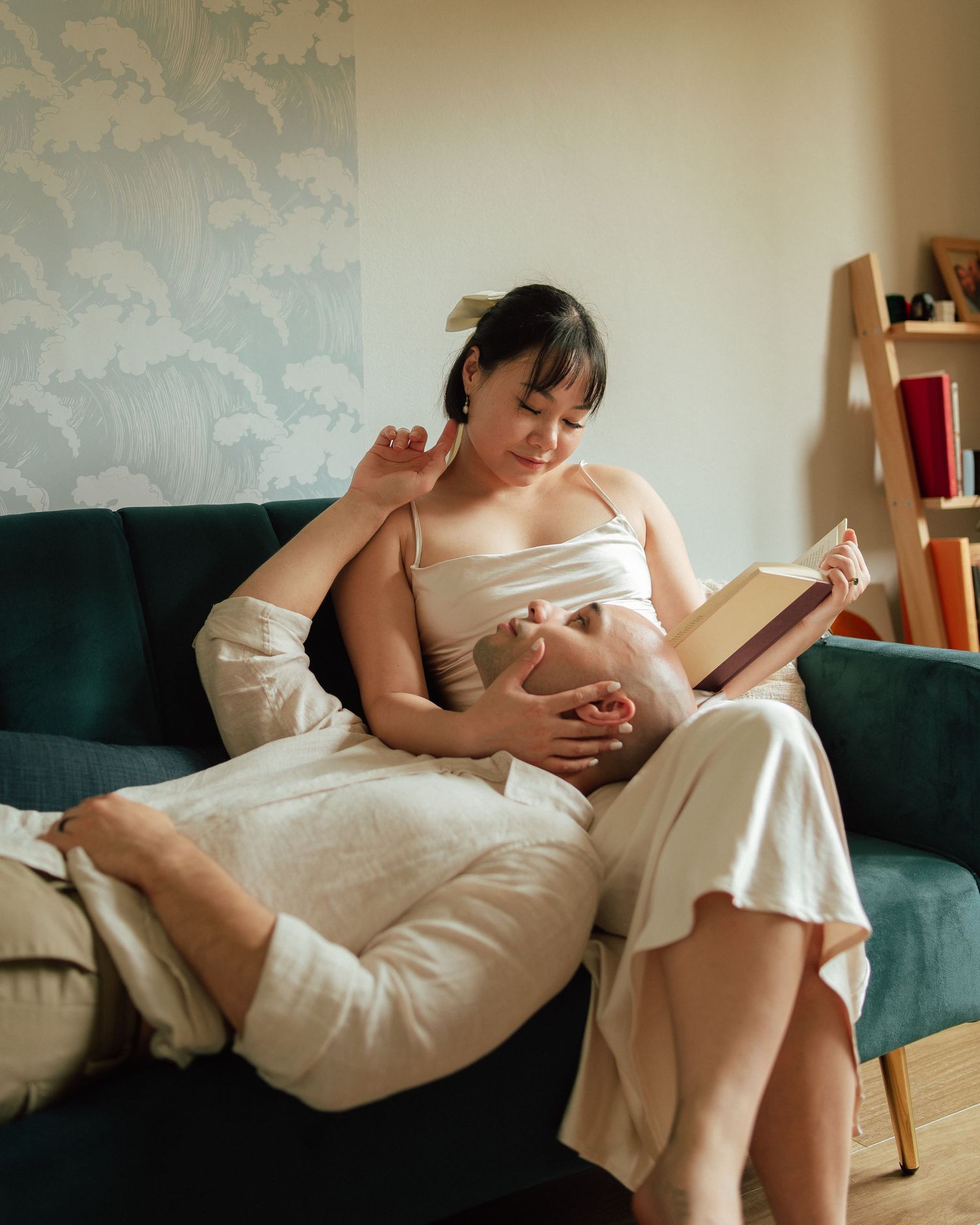 Pregnant woman in white dress reads book on teal sofa while partner rests head on her lap at home.