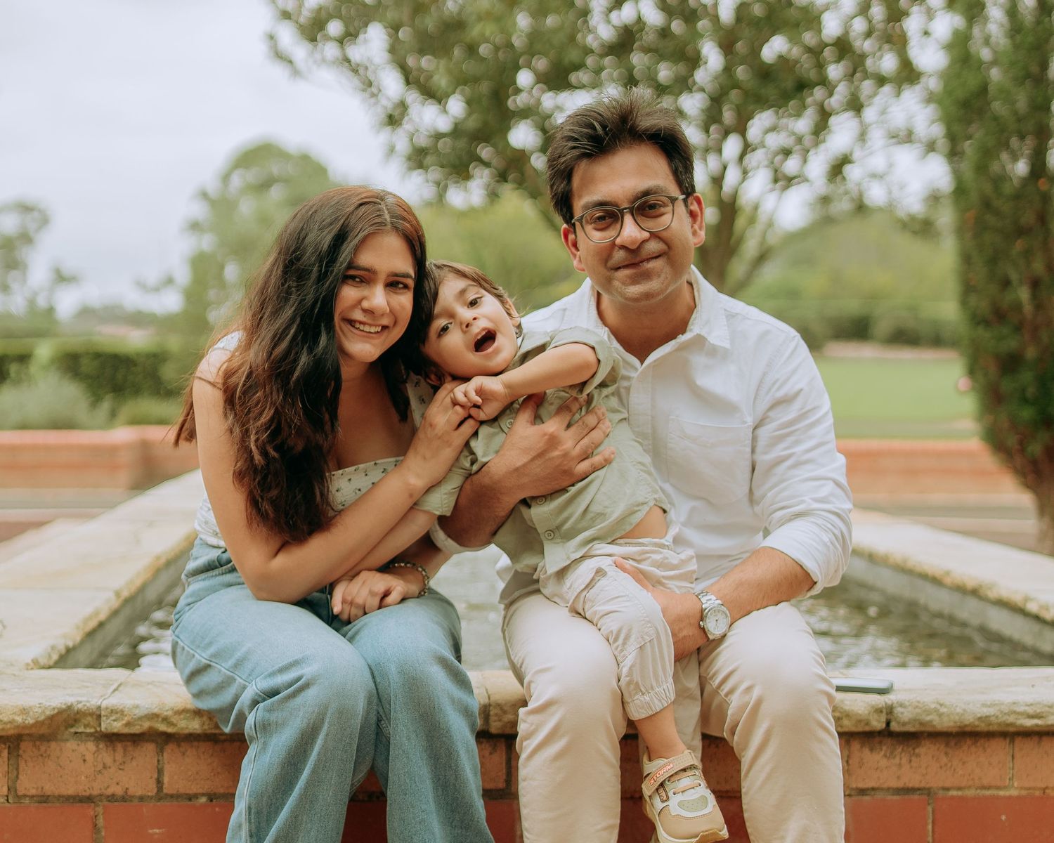 Happy family portrait with smiling parents holding laughing toddler while seated on brick wall in garden setting.