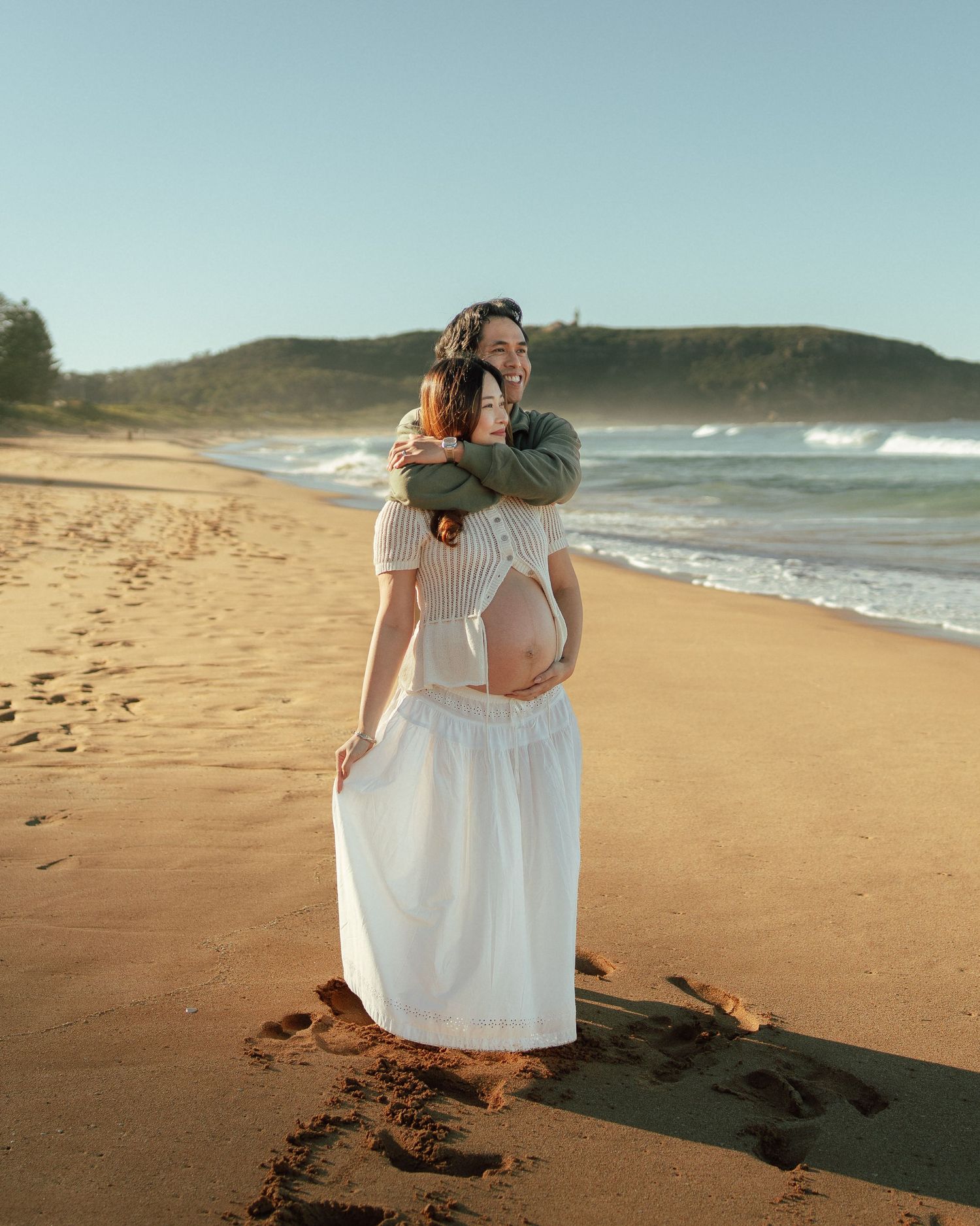 Pregnant woman in white dress receives a warm embrace from partner on a sunny sandy beach.