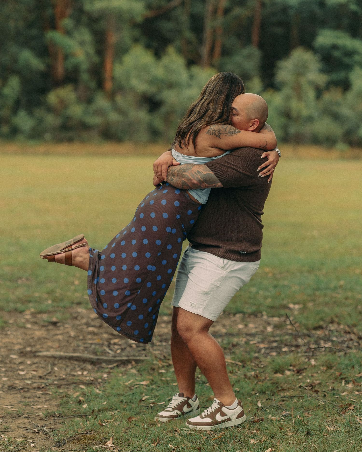 A bald man lifts and embraces his fiancee joyfully in an open green field after a proposal.