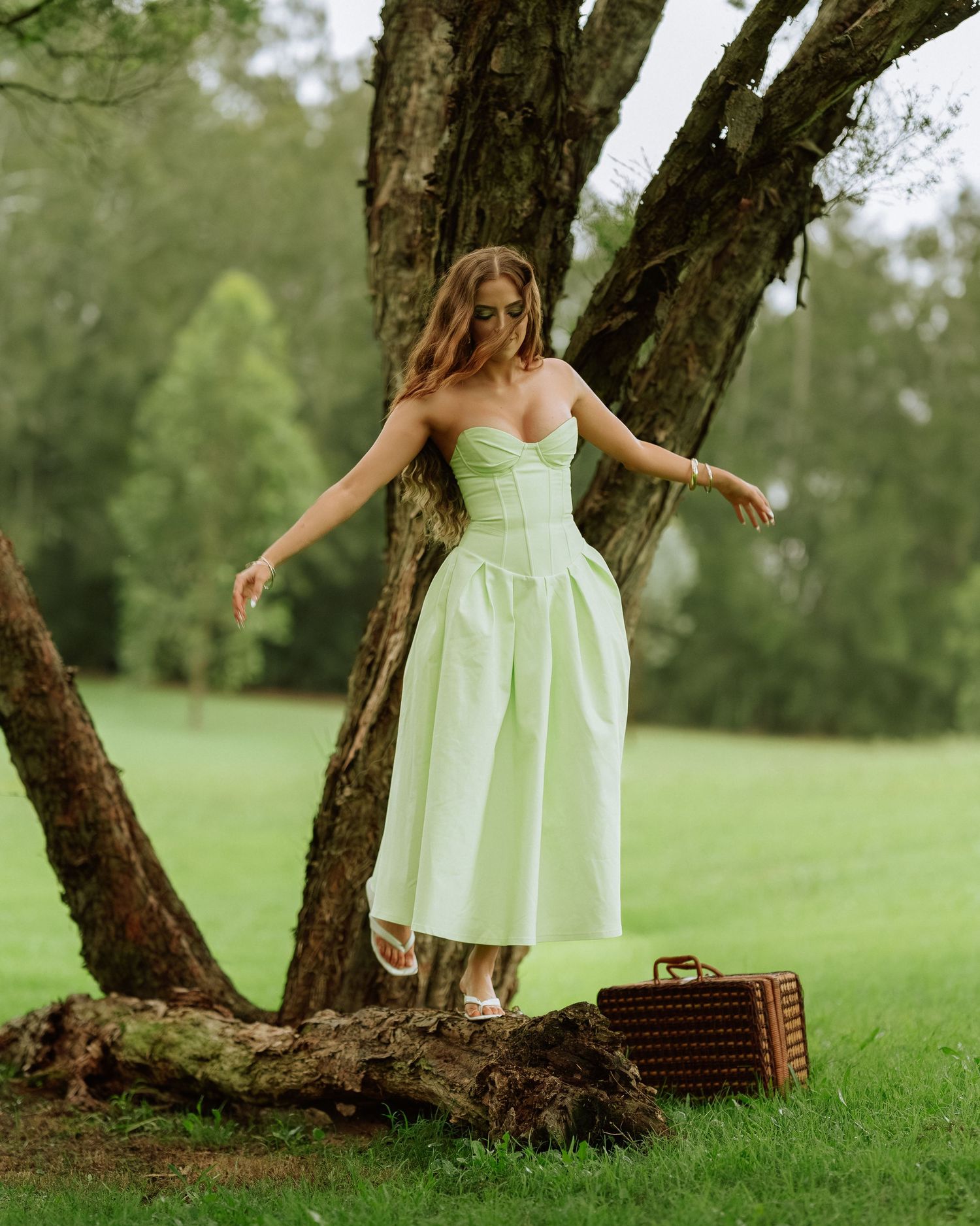 Woman in mint green strapless dress leaning against tree with vintage suitcase in park.