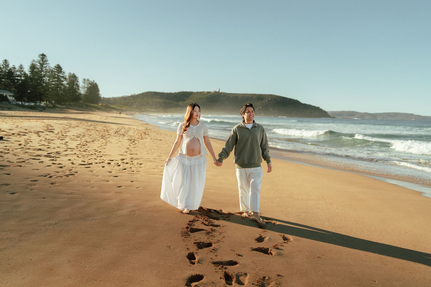 Expecting couple walks hand-in-hand along a sunny sandy beach with ocean waves behind them.